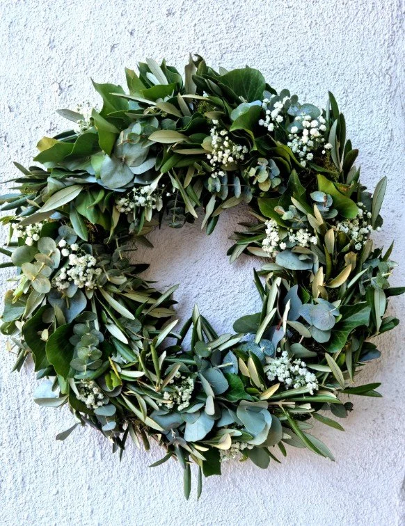 Üppiger, runder Türkranz an weißer Wand, fast ausschließlich aus Blattwerk: blaugraue Eukalyptus-, schmale silbrige Oliven- und glänzend dunkelgrüne Blätter, akzentuiert mit feinen weißen Schleierkraut-Blüten.