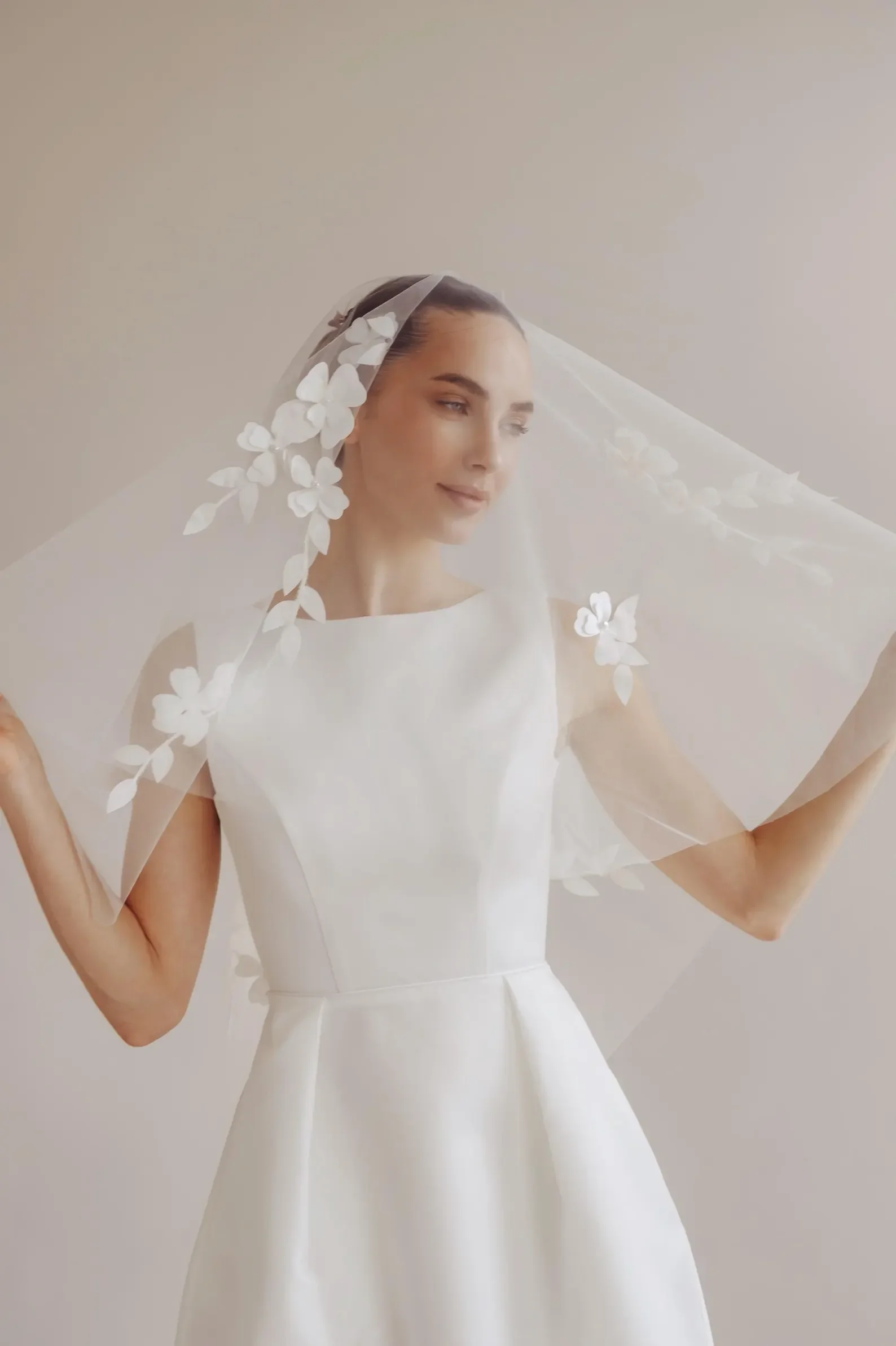 Eine Braut im cleanen, hochgeschlossenen Brautkleid trägt einen modernen Soft-Tüll-Schleier mit floralen 3D-Blütenapplikationen. Inspiration für die aktuellen Brautschleier-Trends 2026 im Holistic Mag.