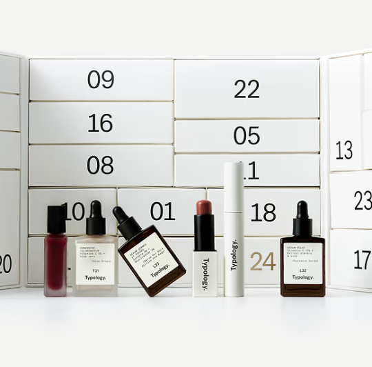 minimalistischer, weißer Beauty-Adventskalender von Typology. Vor den schlichten, nummerierten Schubladen sind ausgewählte Kosmetik- und Hautpflegeprodukte der Marke, auch Seren-Fläschchen mit Pipetten und Lippenprodukte, aufgereiht.
