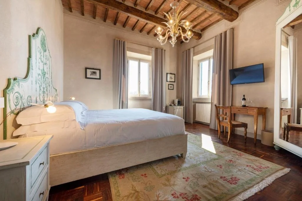 Foto eines hellen Schlafzimmers mit rustikaler Holzbalkendecke, einem eleganten Kronleuchter und einem großen Bett mit kunstvoll grün-weiß verziertem Kopfteil auf einem hellen Blütenteppich über dunklem Holzparkett.