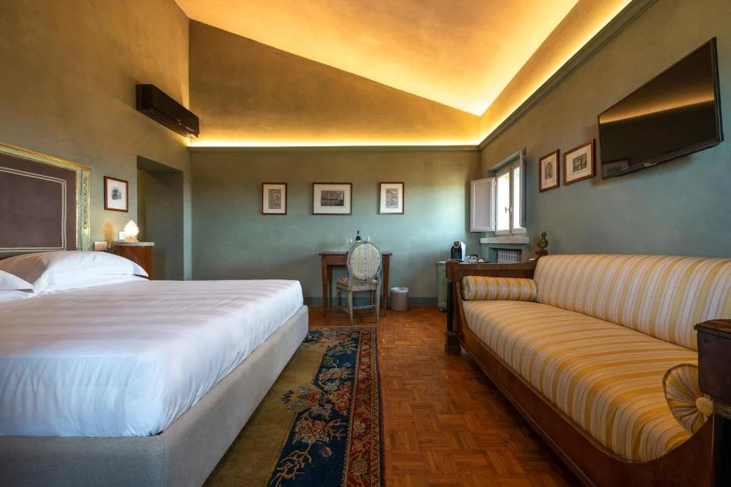 Foto eines eleganten Hotelzimmers mit salbeigrünen Wänden, indirekter Deckenbeleuchtung, einem großen Bett auf einem Orientteppich und einem gelb gestreiften Vintage-Sofa.