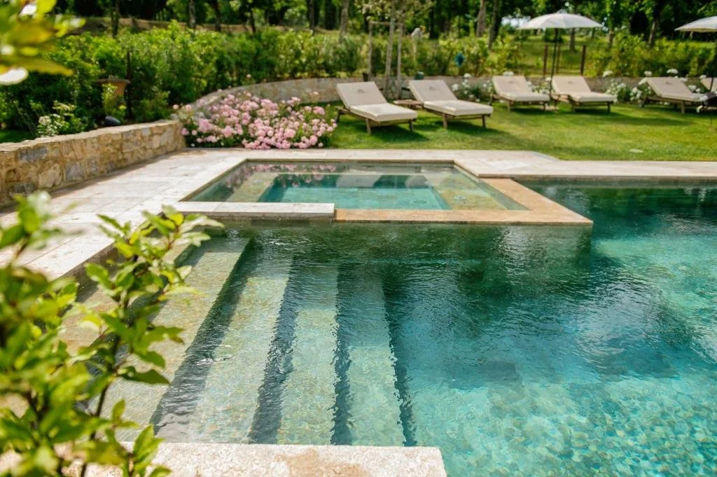 Foto eines Steinpools mit klarem, türkisfarbenem Wasser und breiten Einstiegsstufen. Im Hintergrund grenzt eine grüne Wiese mit weißen Sonnenliegen, Sonnenschirmen und blühenden rosa Rosensträuchern an.