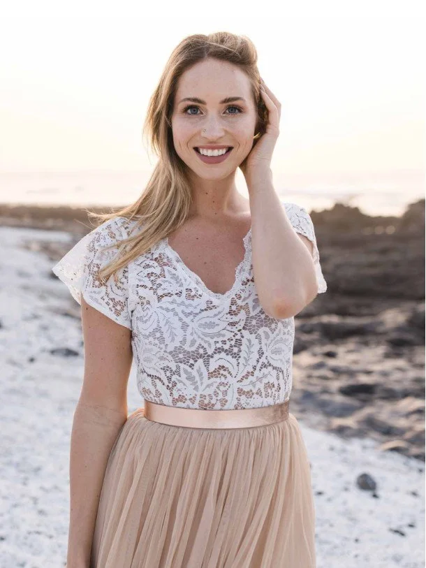 Boho-Braut am Strand bei Sonnenuntergang in einem Zweiteiler mit kurzärmeligem Spitzentop mit V-Ausschnitt, einem Roségold-Gürtel und einem nudefarbenen Tüllrock.