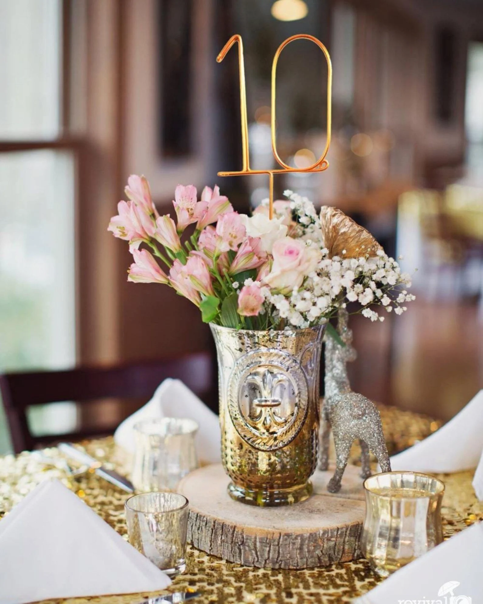 Eine filigrane, goldene Drahtzahl „10“, die als Stecker in einer prachtvollen Blumenvase auf einem mit Goldpailletten verzierten Tisch steht.