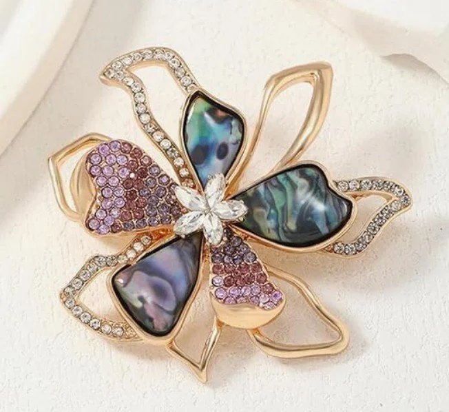 Goldene, blütenförmige Brosche auf Weiß: zwei Blütenblätter mit dunklem Perlmutt, zwei mit lila Kristallen, mittig ein kleiner Kristallstern. Edler Broschenschmuck für elegante Outfits.