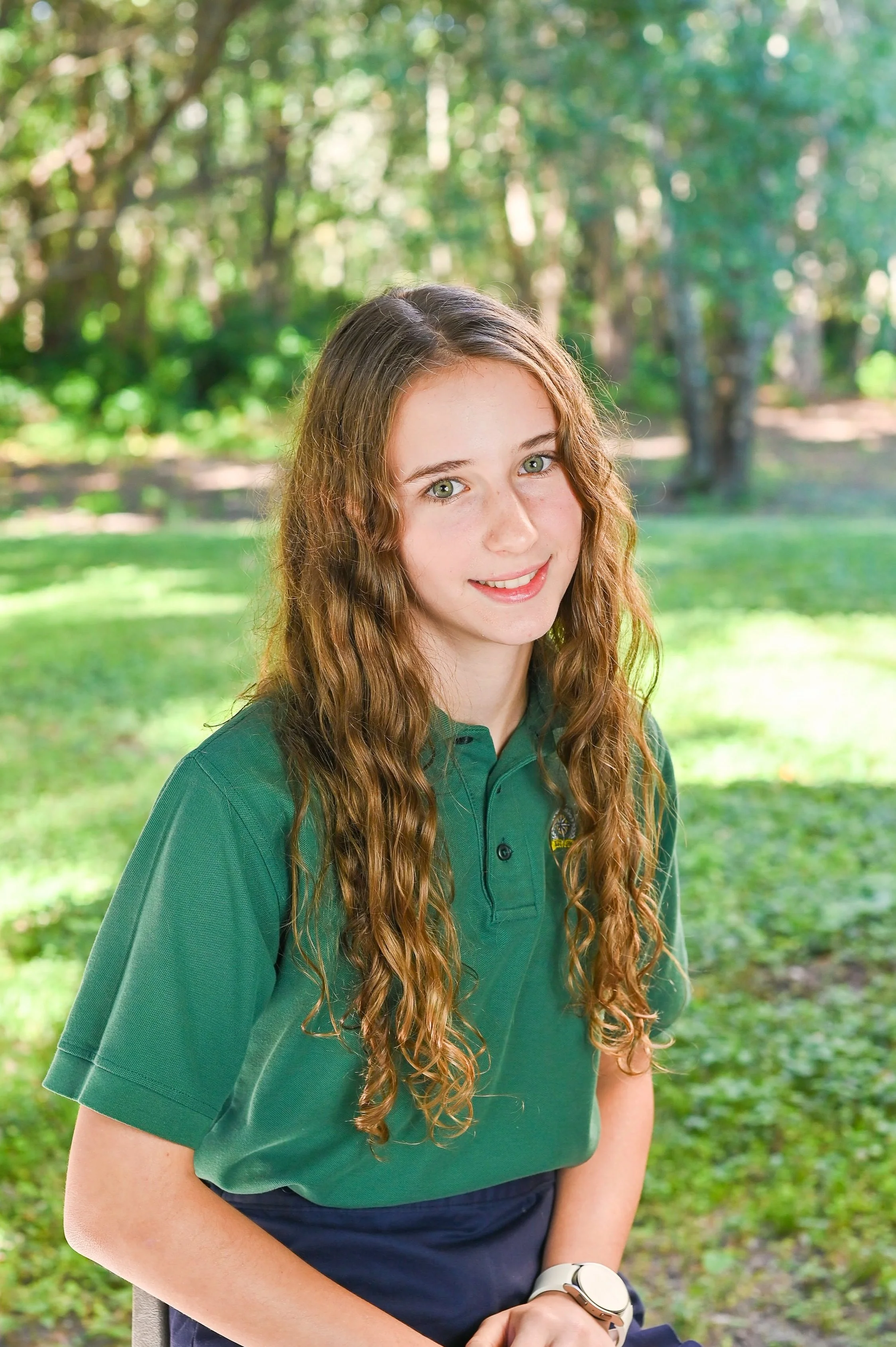 A young woman with long, curly brown hair, wearing a green polo shirt and dark pants, sitting outdoors in a park with lush green grass and trees in the background.