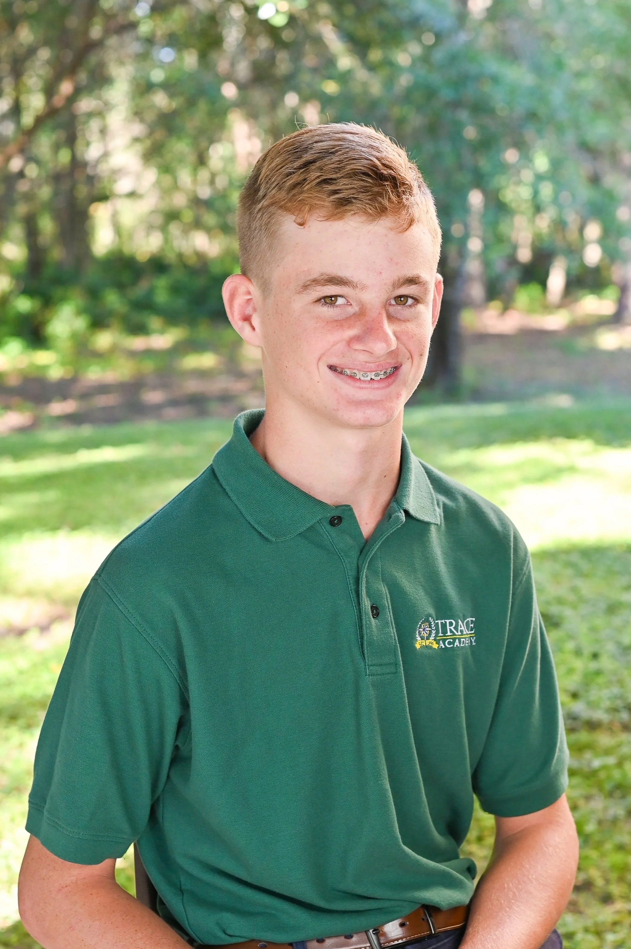 A young man with short red hair, wearing braces, smiling, and dressed in a green polo shirt with a logo that reads "Trace Academy," standing outdoors in a park with trees and sunlight in the background.