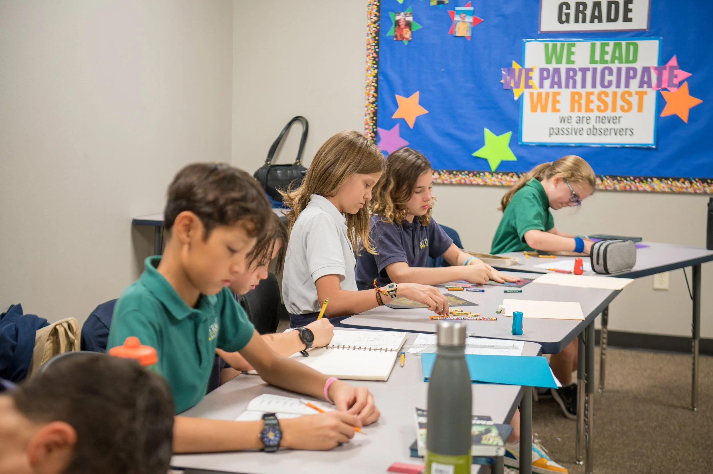Students sitting at desks taking notes in a classroom with a colorful bulletin board that has motivational phrases and stars.