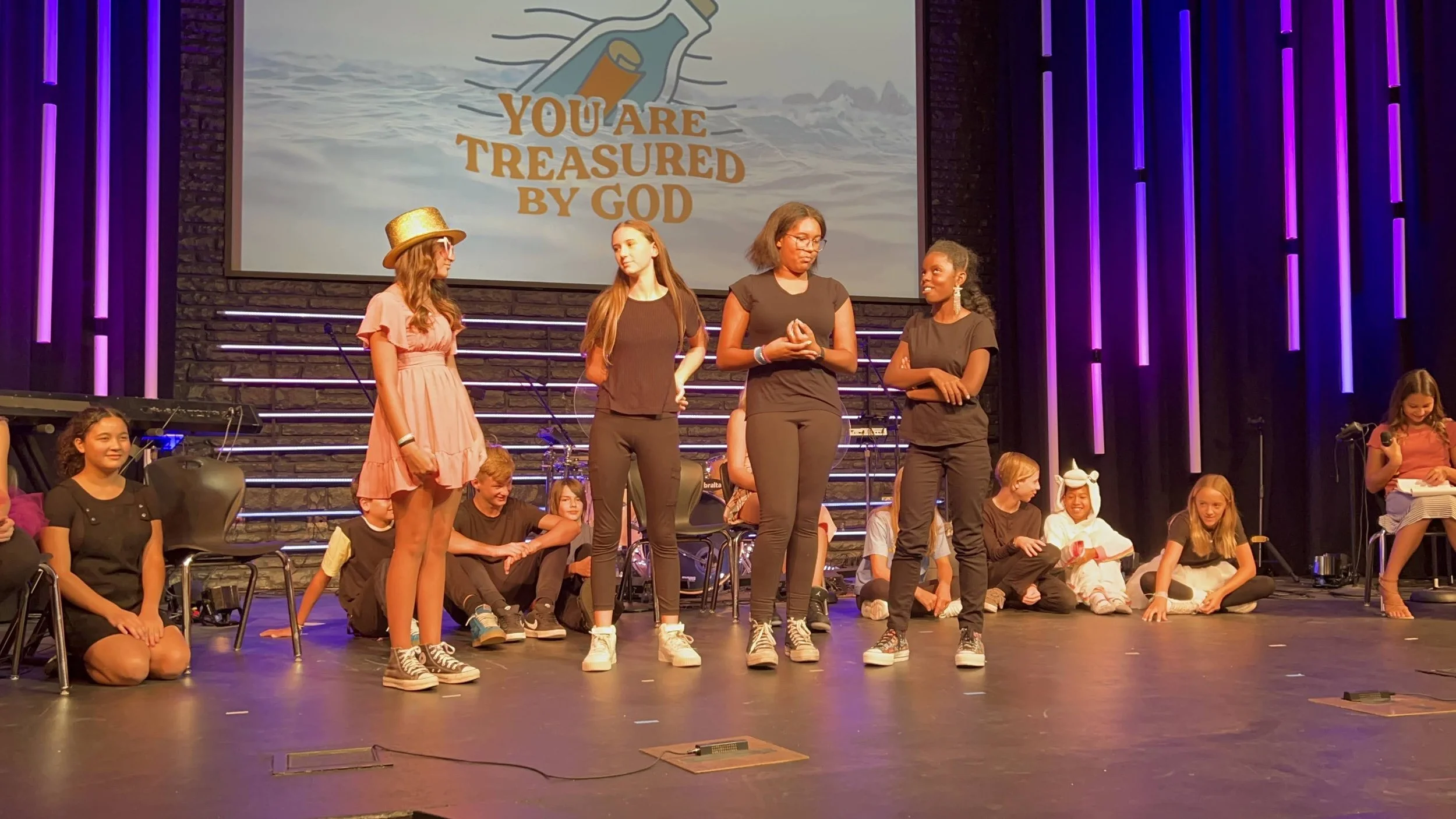 Children and teenagers on stage during a performance or presentation, with a large screen behind them displaying the message 'You are treasured by God' in colorful text.