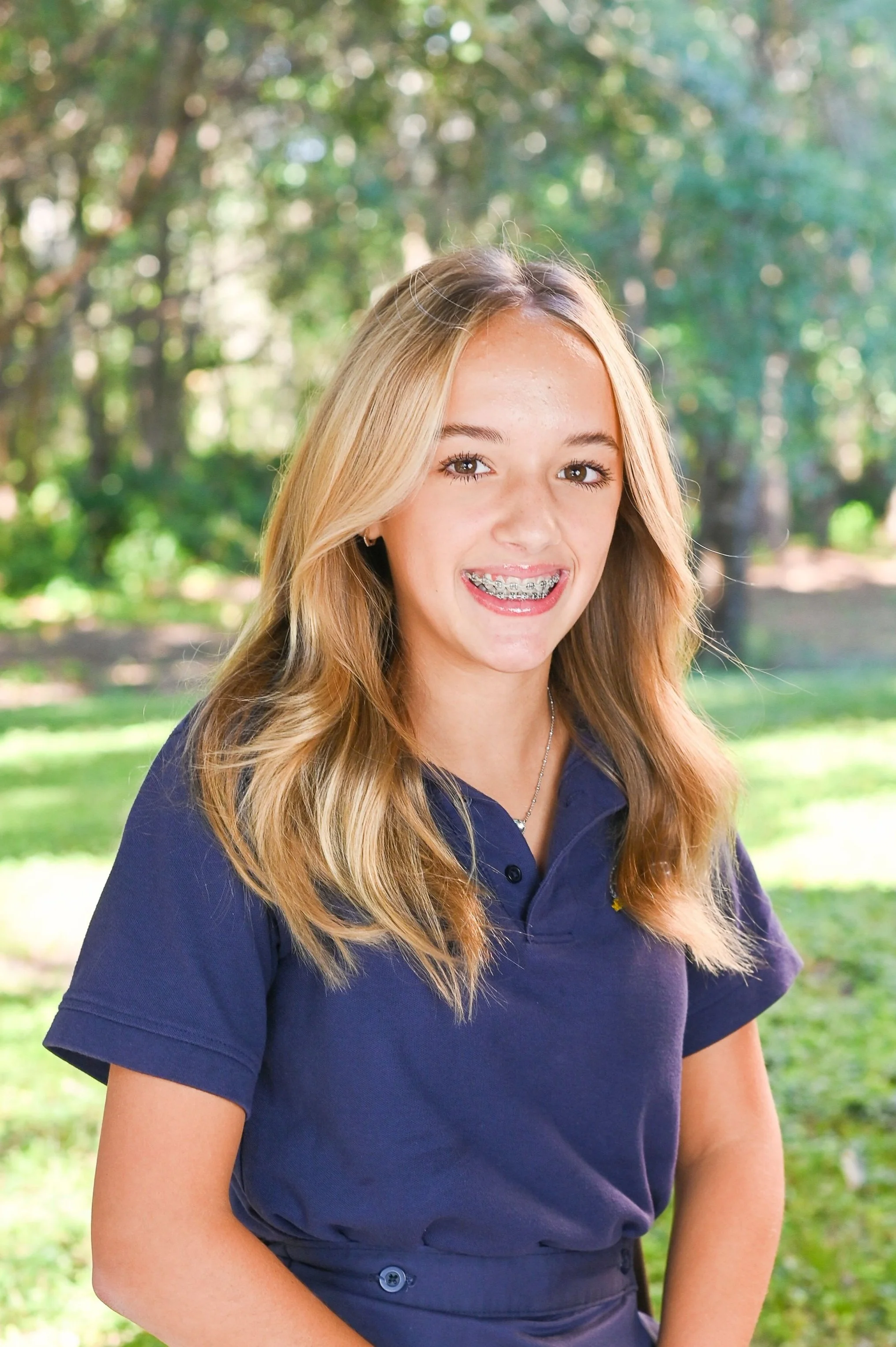 A teenage girl with long blonde hair, braces, and a necklace, smiling outdoors in a park with green trees and sunlight in the background.