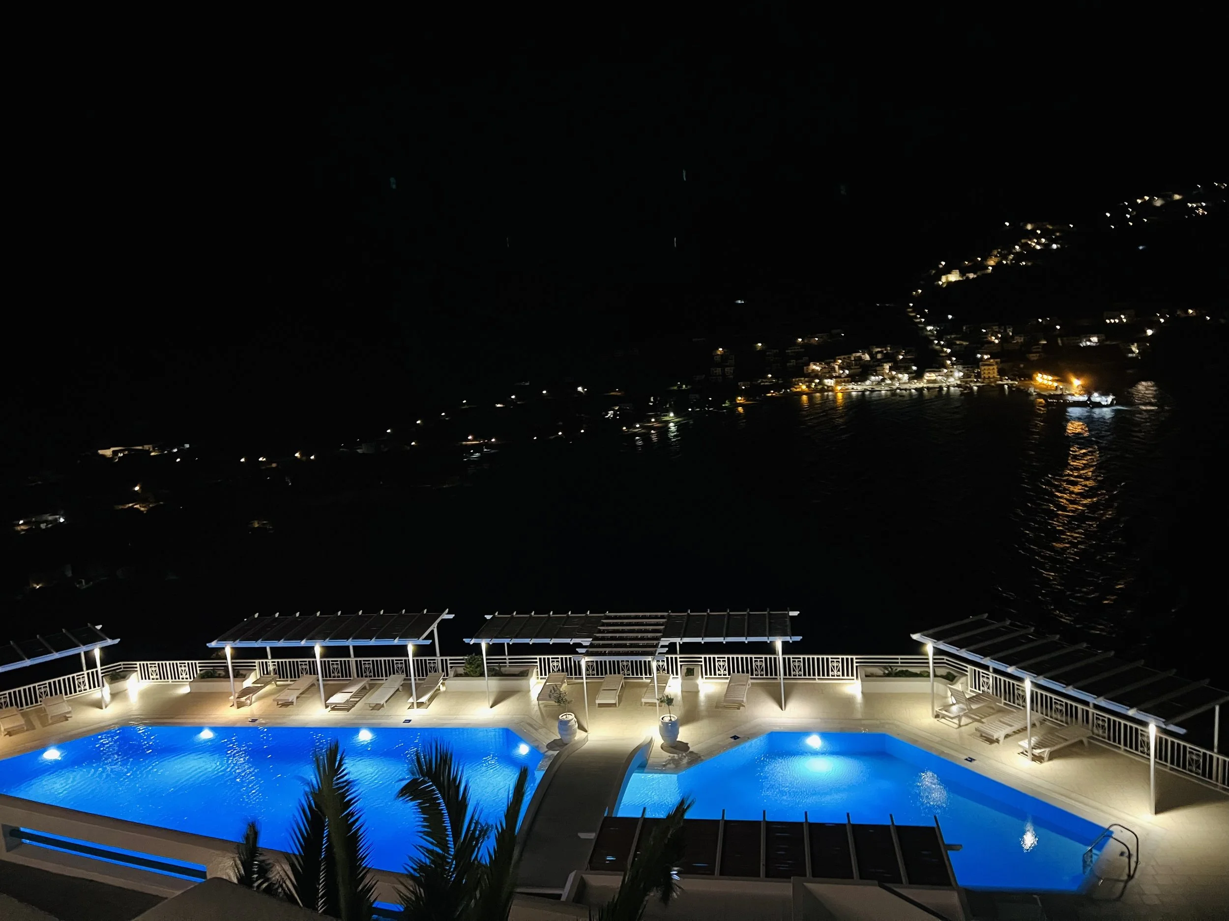 Nighttime view of a lit swimming pool on a balcony overlooking a dark ocean with distant lights from a coastal town.