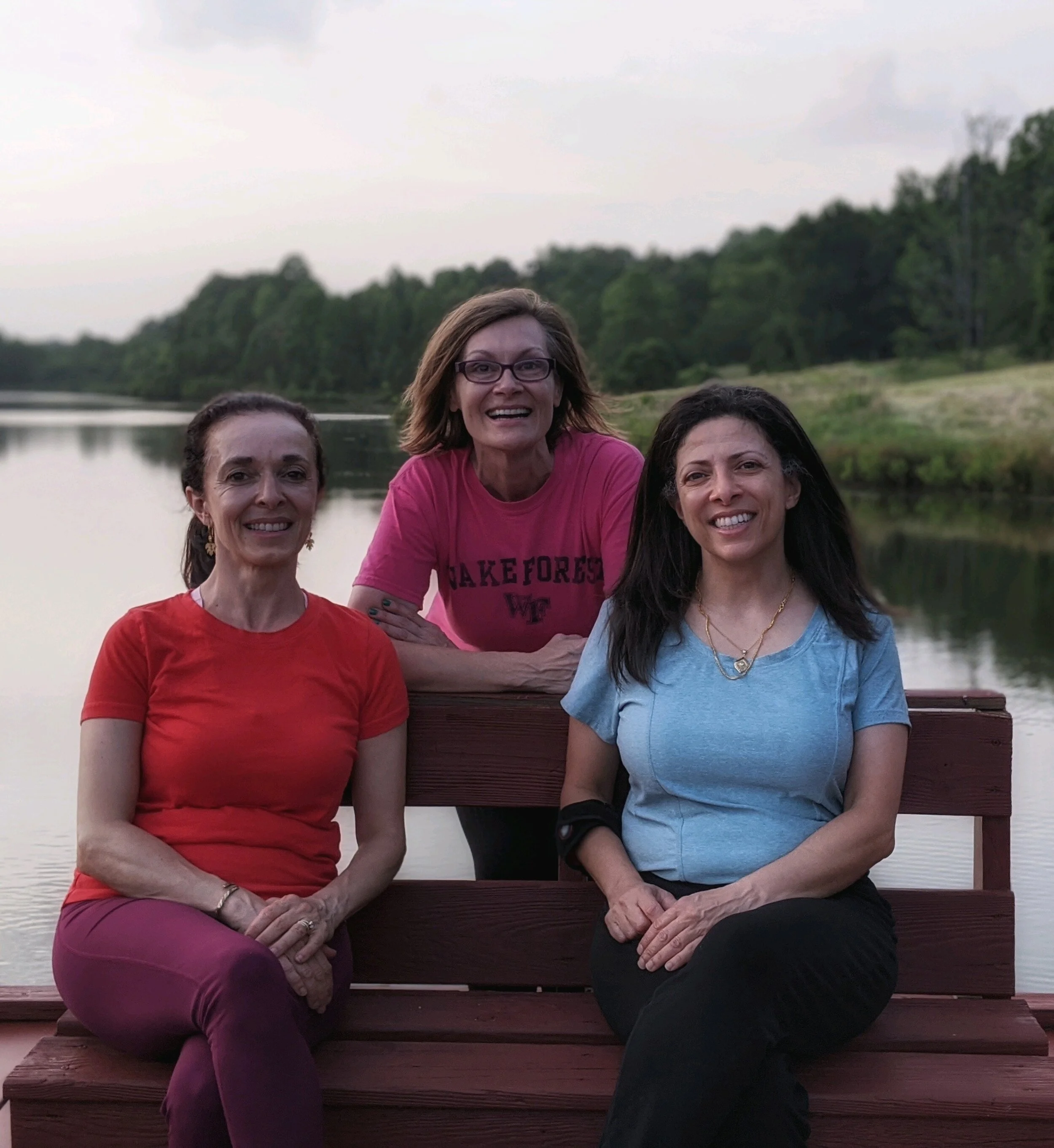 Three women sitting on a wooden bench by a lake, smiling at the camera. The woman in the middle is wearing glasses and a pink shirt, while the other two women are wearing red and blue shirts.