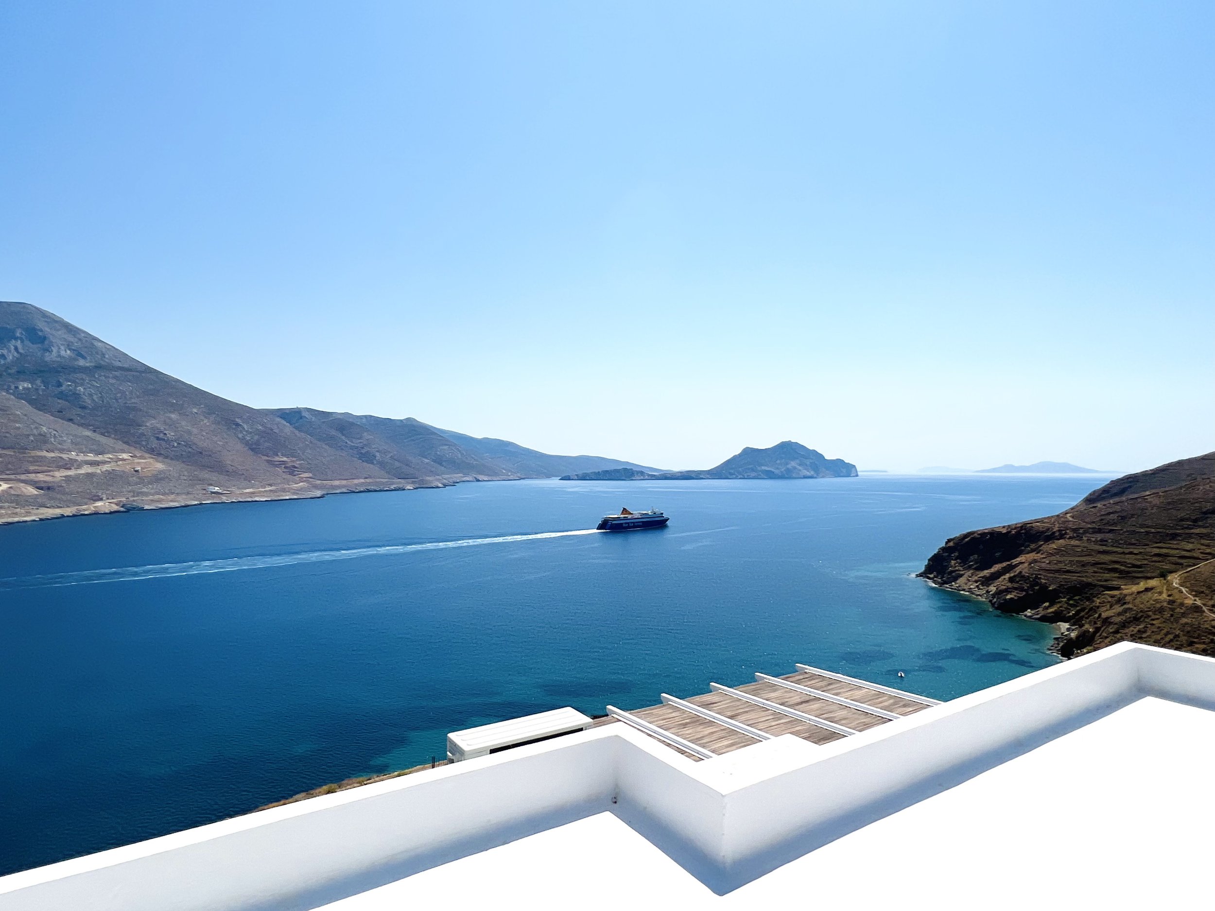 View of a large body of water with a ferry boat sailing across, surrounded by hills and mountains under a clear blue sky, with part of a white building or balcony in the foreground.