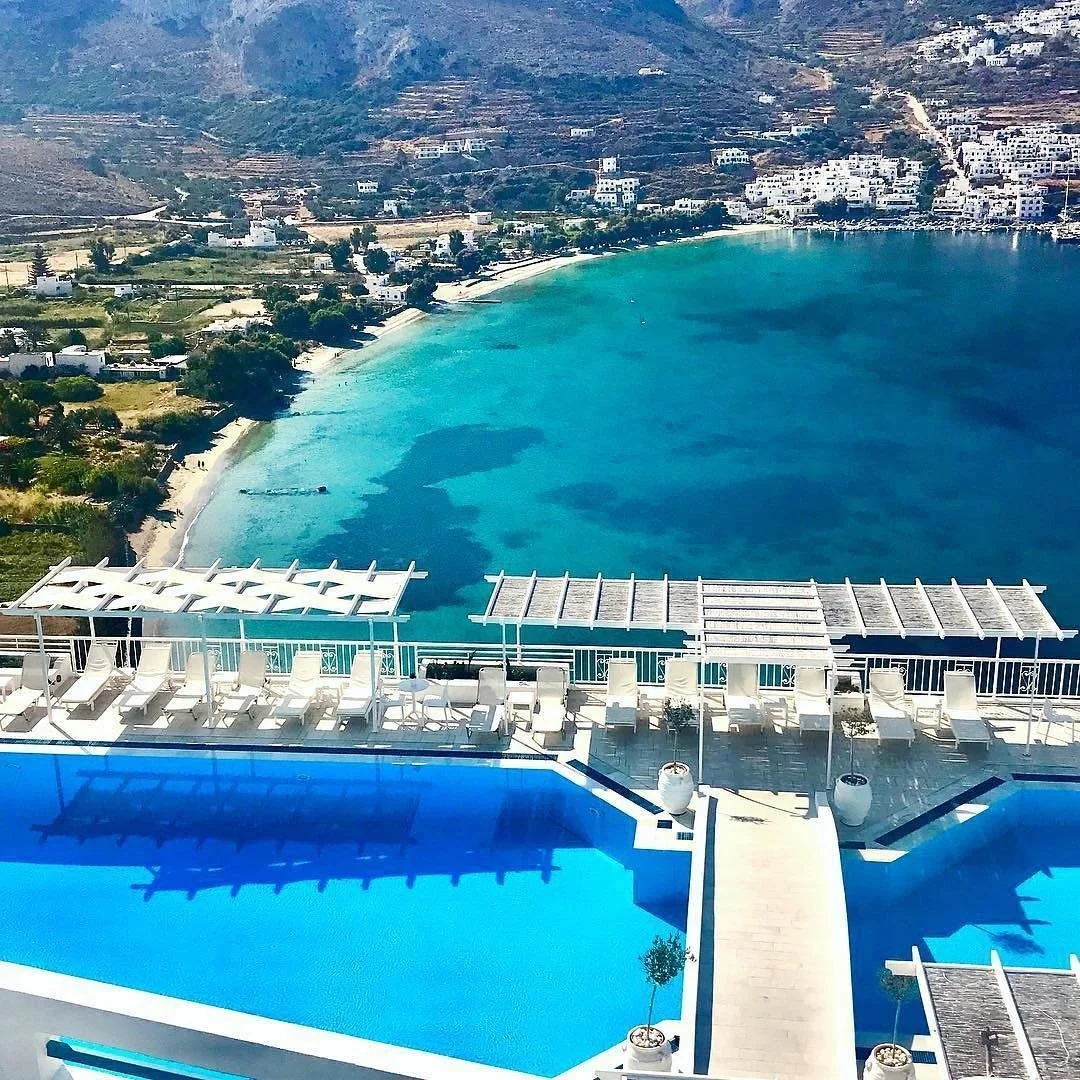View of a resort with a swimming pool overlooking a turquoise bay, with white buildings and hills in the background.
