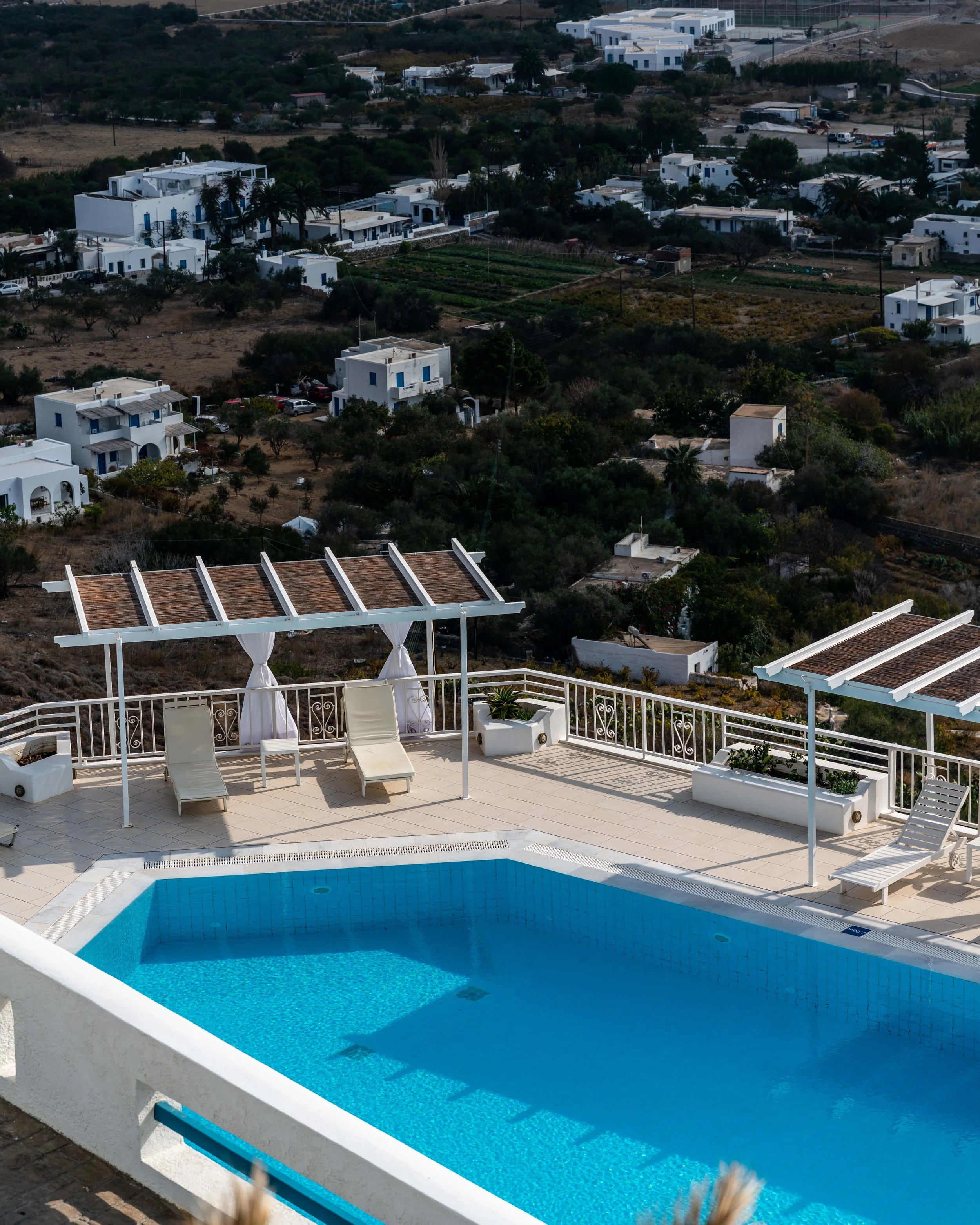 A rooftop swimming pool with lounge chairs and shaded cabanas overlooking a scenic hillside village with white buildings and lush greenery.