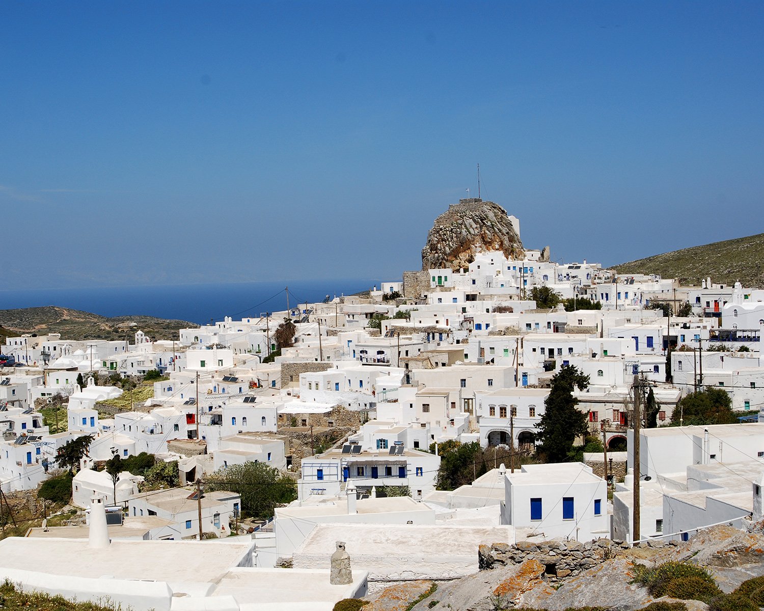 White buildings with blue accents on a hillside overlooking the sea, with a rocky hill in the background under a clear blue sky.