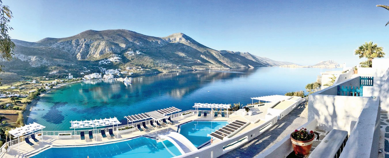 A scenic coastal landscape with mountains, a body of water, and white buildings with outdoor pools and lounge chairs in the foreground.