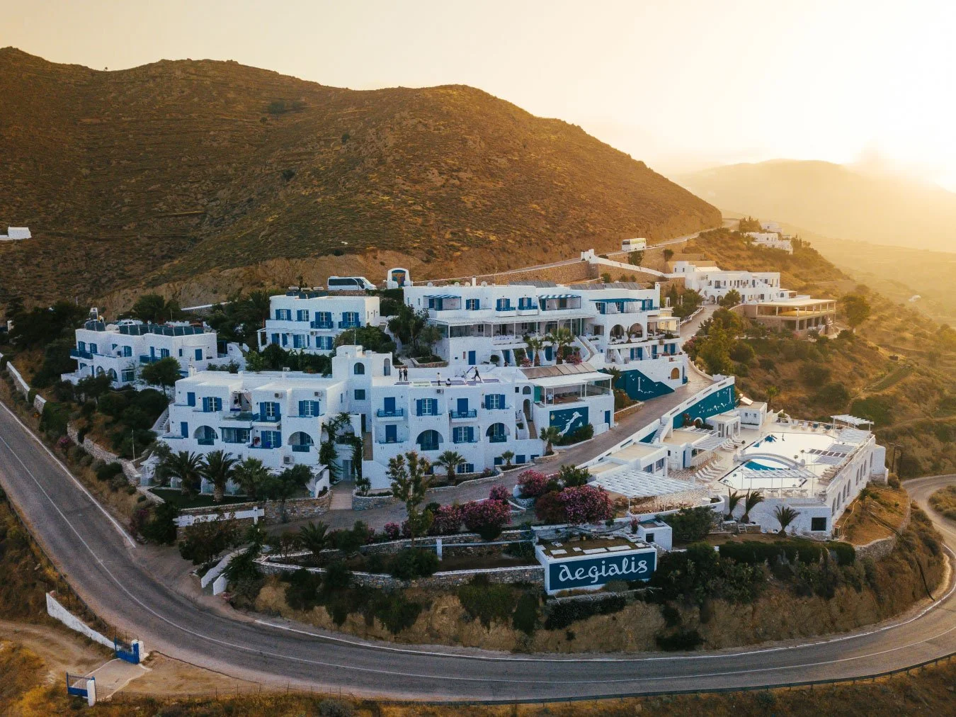 White Greek-style hotel with blue accents on a hillside at sunset, featuring multiple levels, balconies, swimming pools, and a sign that says 'Aegialis' near the road.