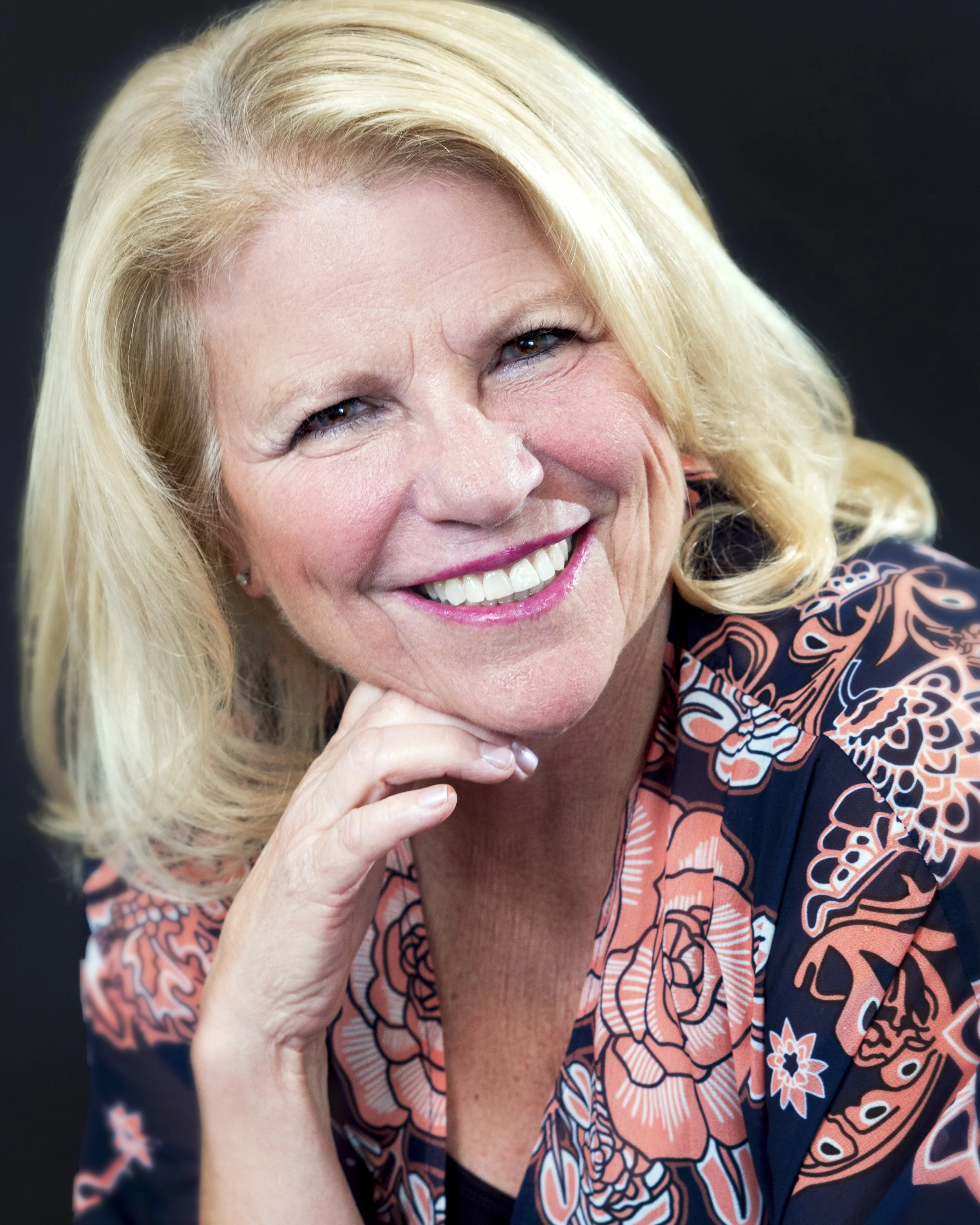 Headshot of lynne in floral shirt, smiling with blond hair. Her chin sits on her fingers.