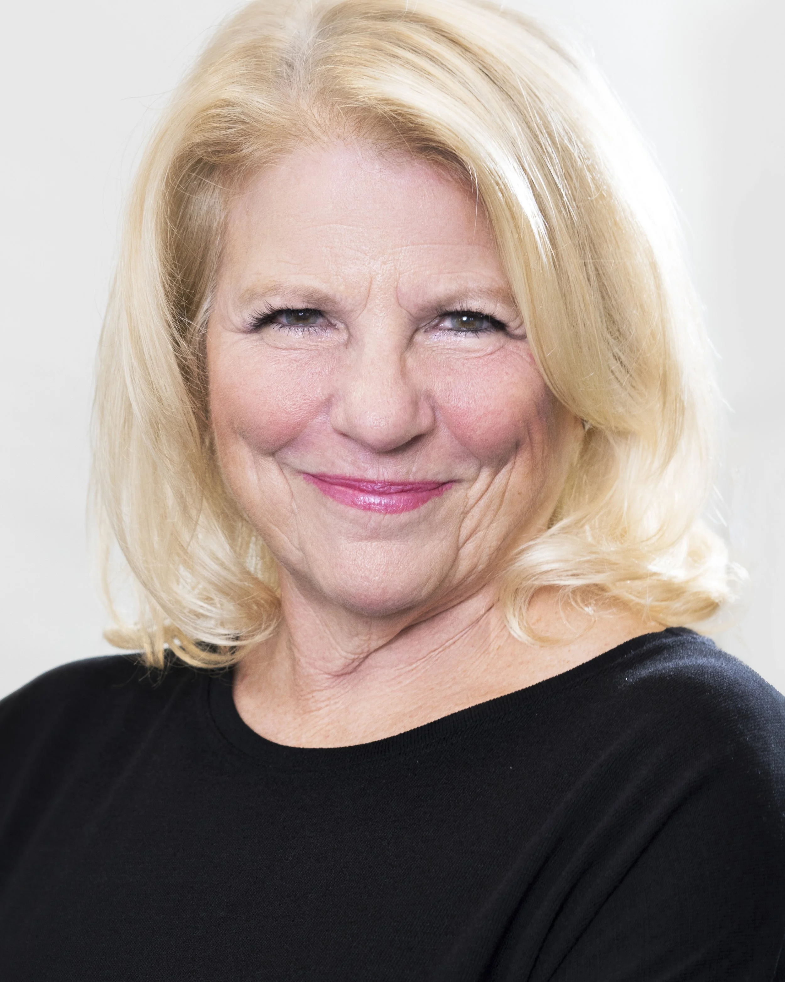 Headshot of Lynne. Blond hair, facing left, and smiling at camera in a black shirt.