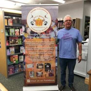 Man standing next to a banner in a library, wearing a purple "END HUNGER" t-shirt. The banner promotes a community food initiative called "Bill's Breads and Fresh Bread" and includes images and text about feeding the hungry. Bookshelves are visible in the background.