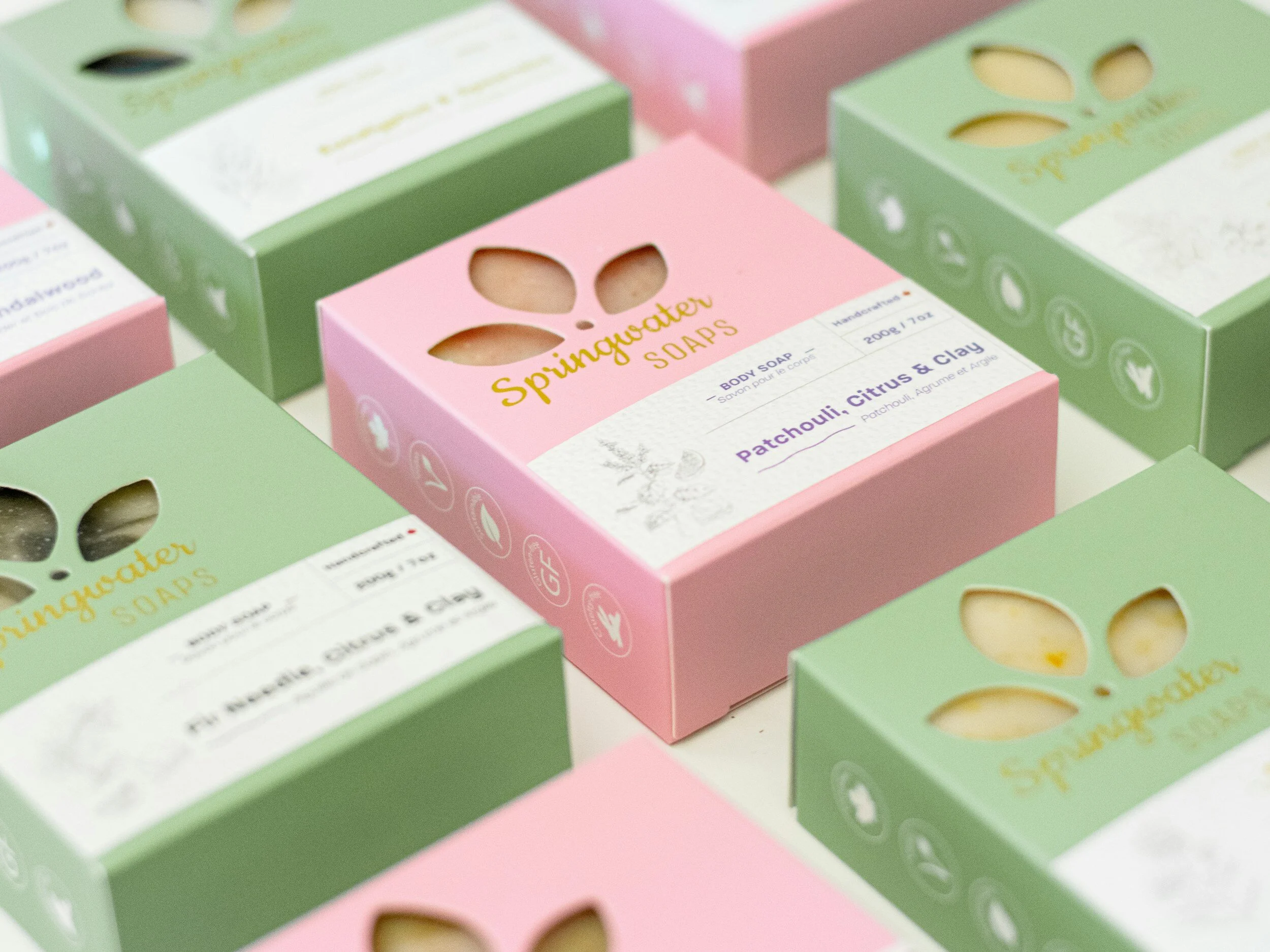 Various boxes of Springwater Soaps in pink and green packaging, featuring scents like Patchouli, Citrus & Clay, and Fir Needle, Citrus & Clay.