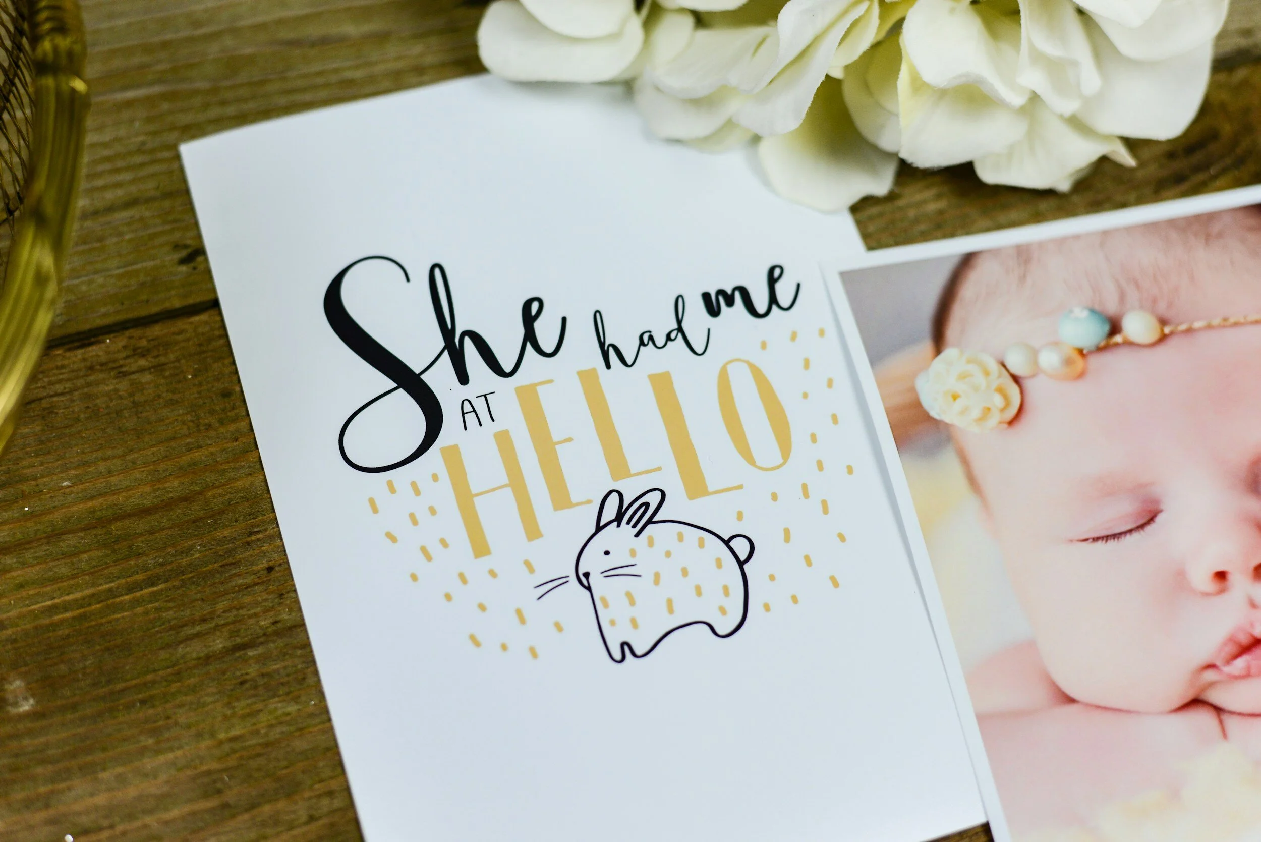 Greeting card with the message "She had me at hello" featuring a cute bunny illustration, next to a photo of a sleeping baby with a floral headband, placed on a wooden surface with white flowers.