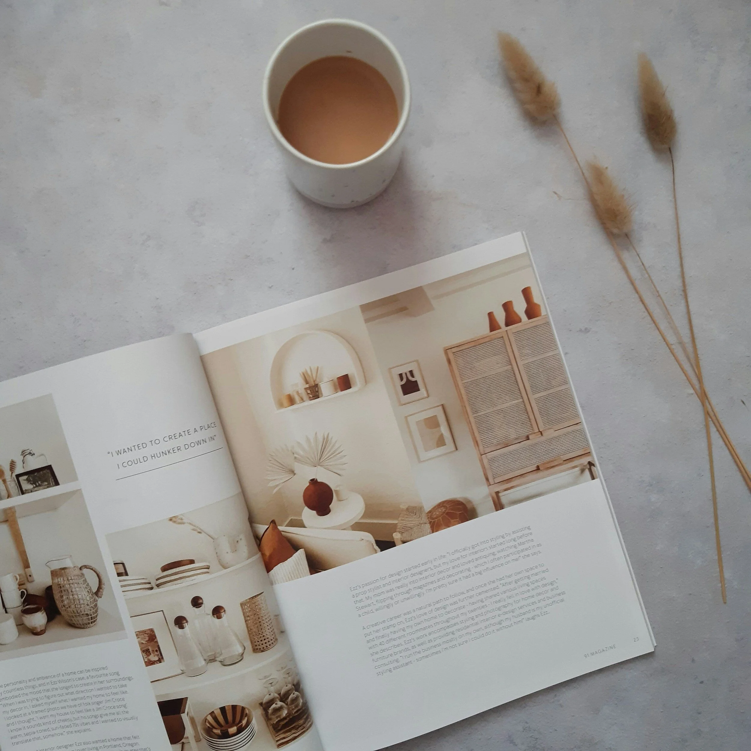 Open magazine with interior design photos, a cup of coffee on a light surface, and dried decorative grass.