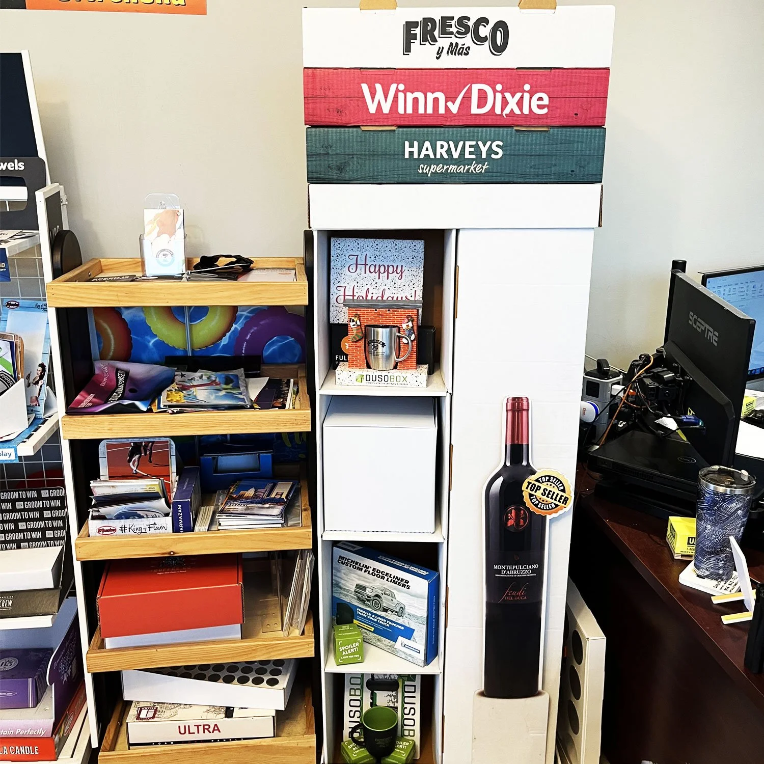 Office corner with shelves and wine display, featuring signage for Fresco y Más, Winn-Dixie, and Harveys supermarket, along with miscellaneous office supplies and decorations.