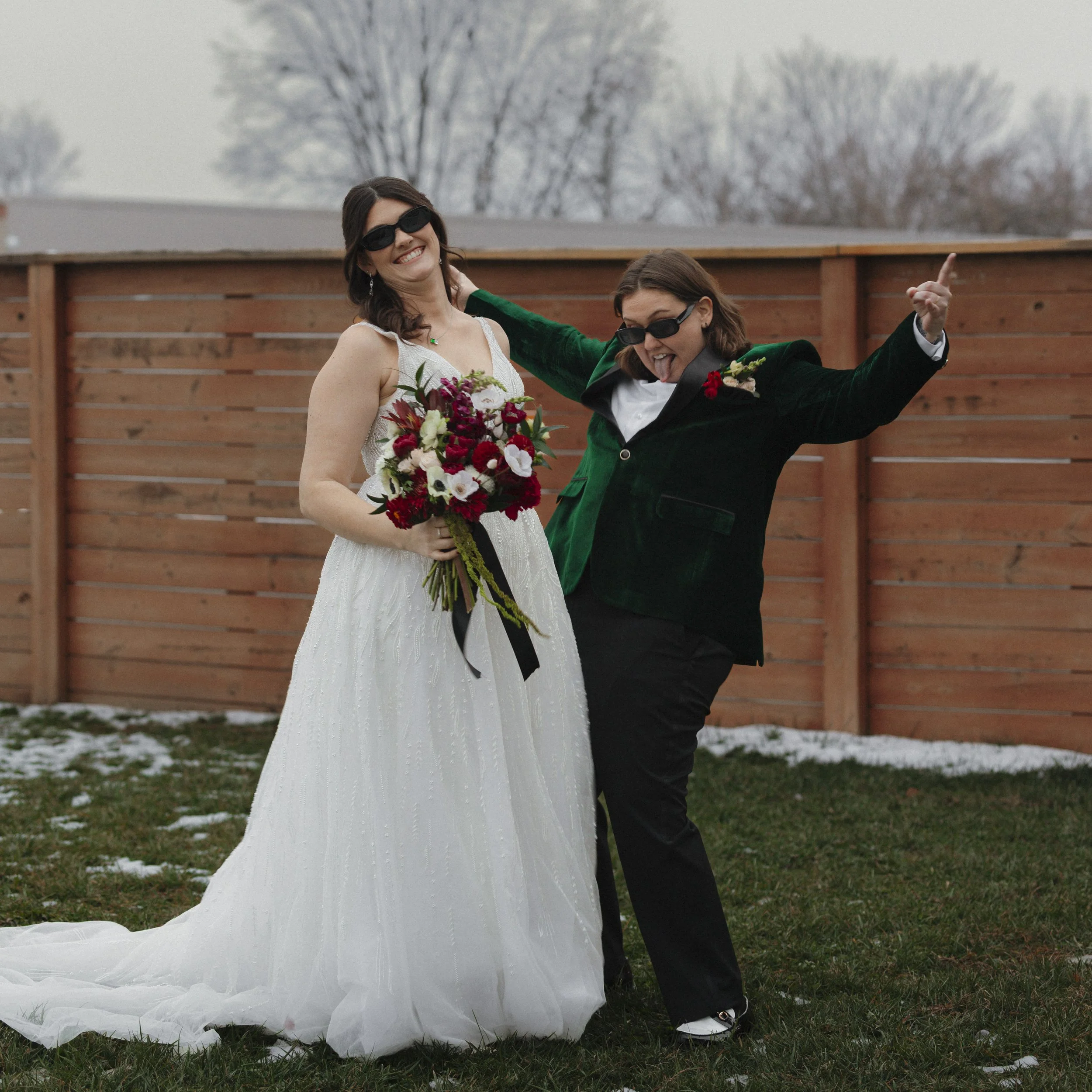 Two women, one in a white wedding dress holding a bouquet, and the other in a dark green jacket, posing outdoors in front of a wooden fence on a chilly day.