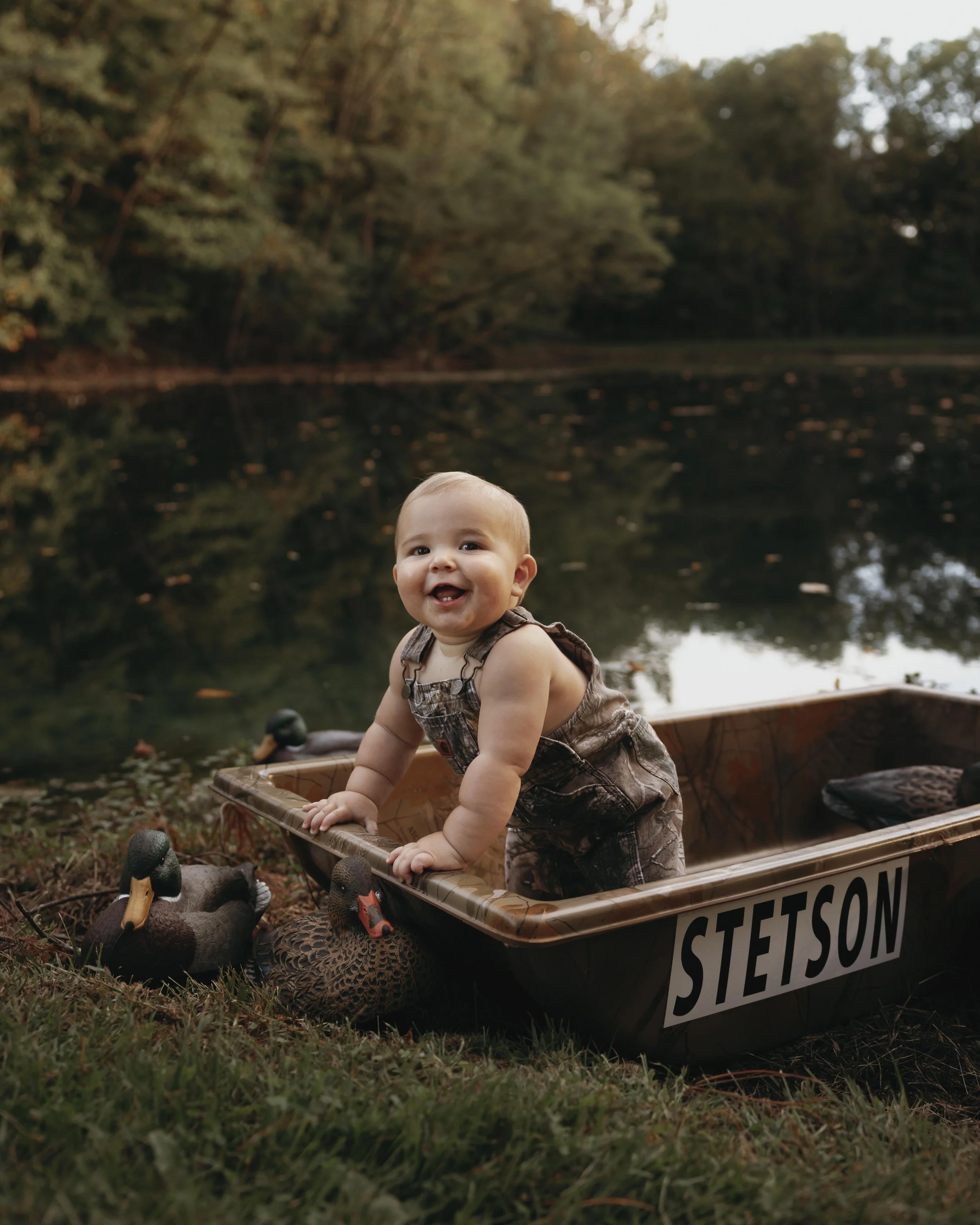 A smiling baby wearing overalls, standing in a camouflage boat named Stetson, outdoors near a lake with trees in the background, accompanied by two duck decoys on the ground.