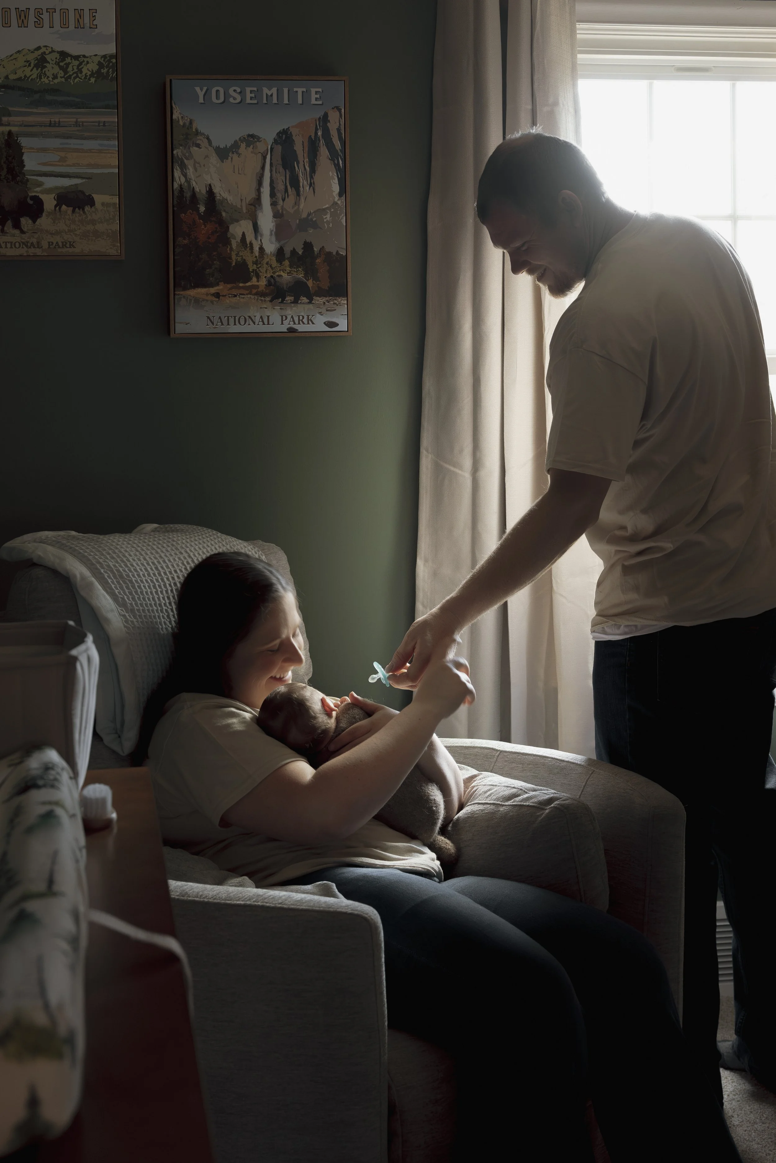A woman sitting on a chair holding a newborn baby and smiling at him, while a man standing next to her feeds the baby with a spoon in a warmly lit room.