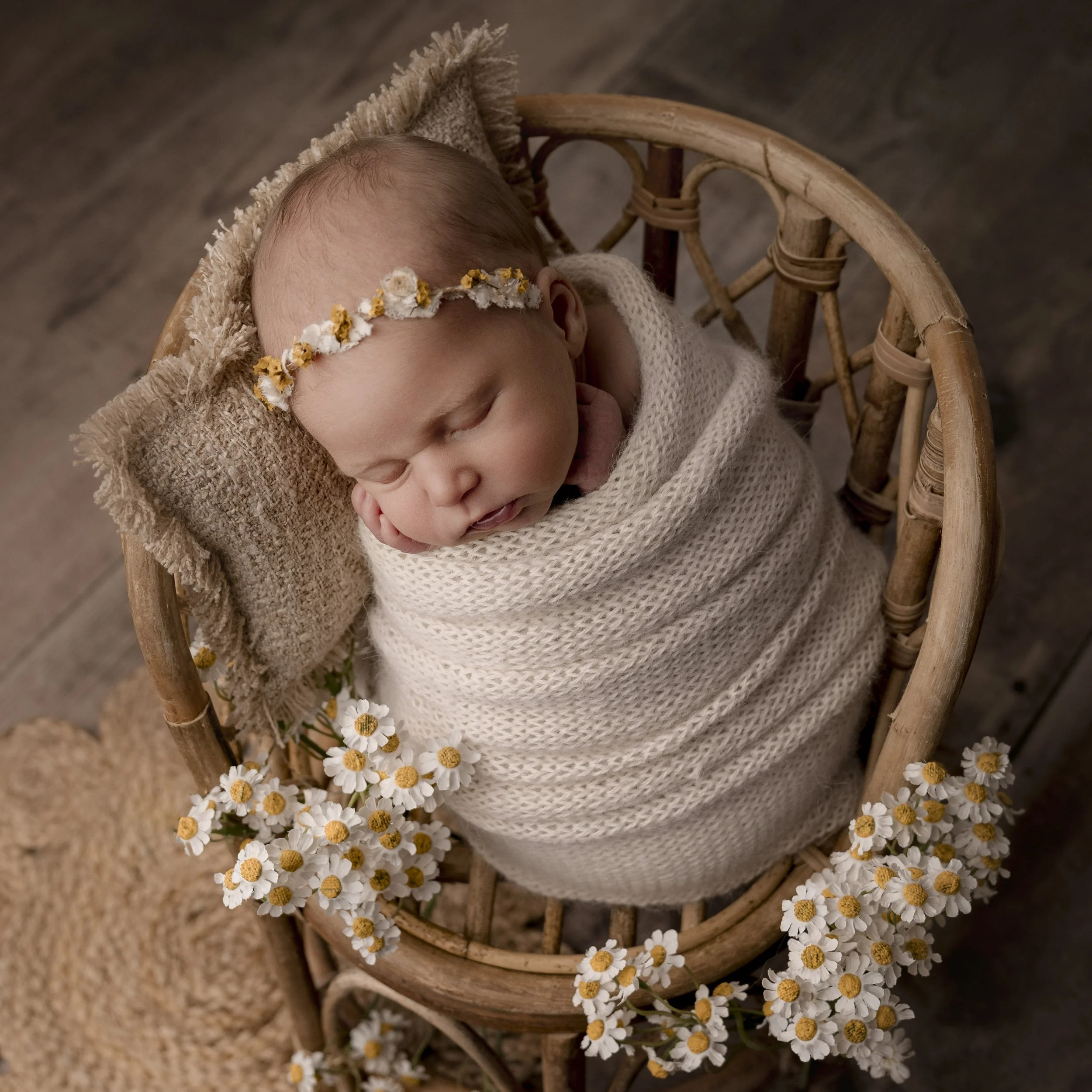 A sleeping baby with a floral headband, swaddled in a cream blanket, lying in a wicker basket decorated with daisies.