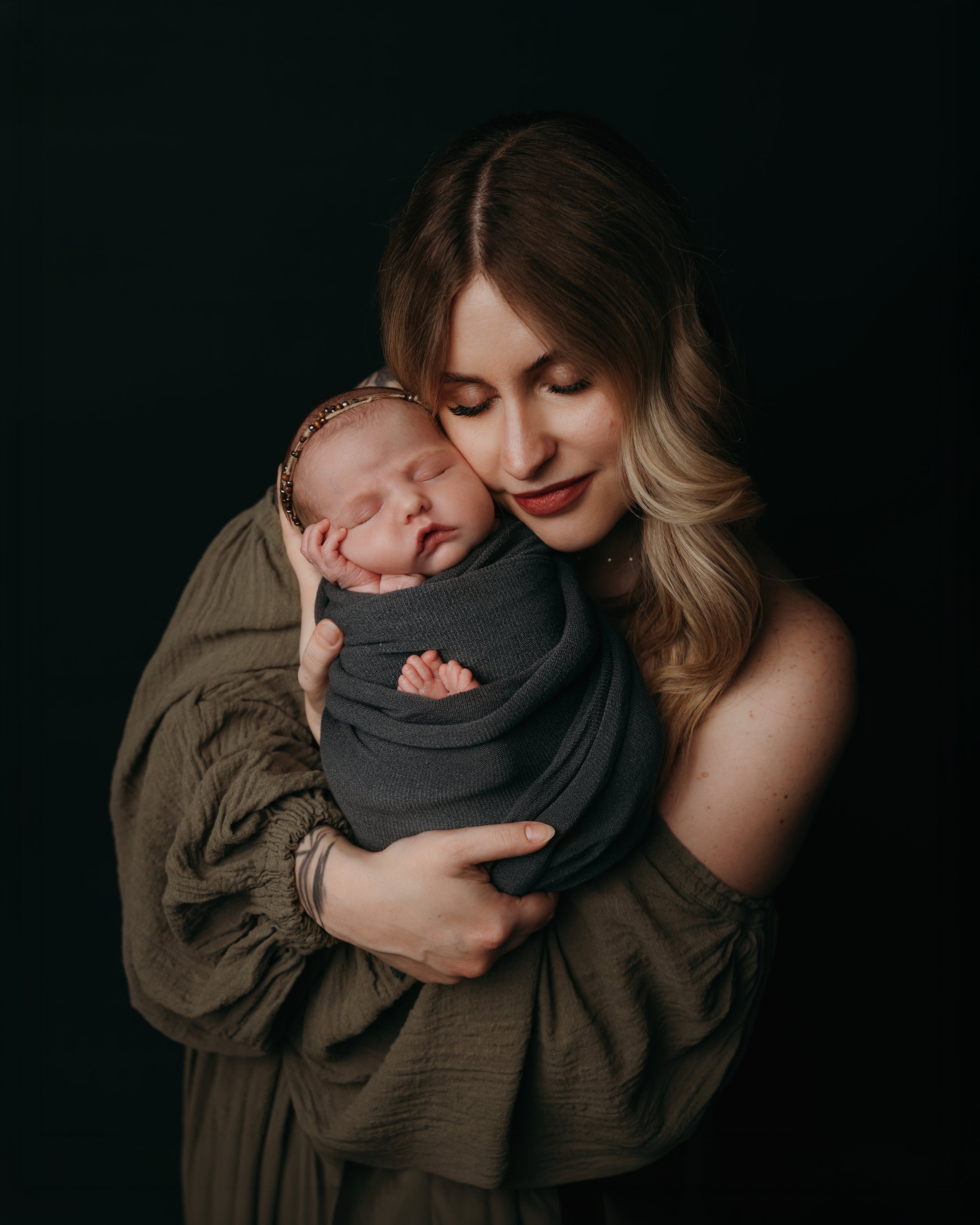 A woman holding a sleeping baby wrapped in a dark gray blanket against a black background.