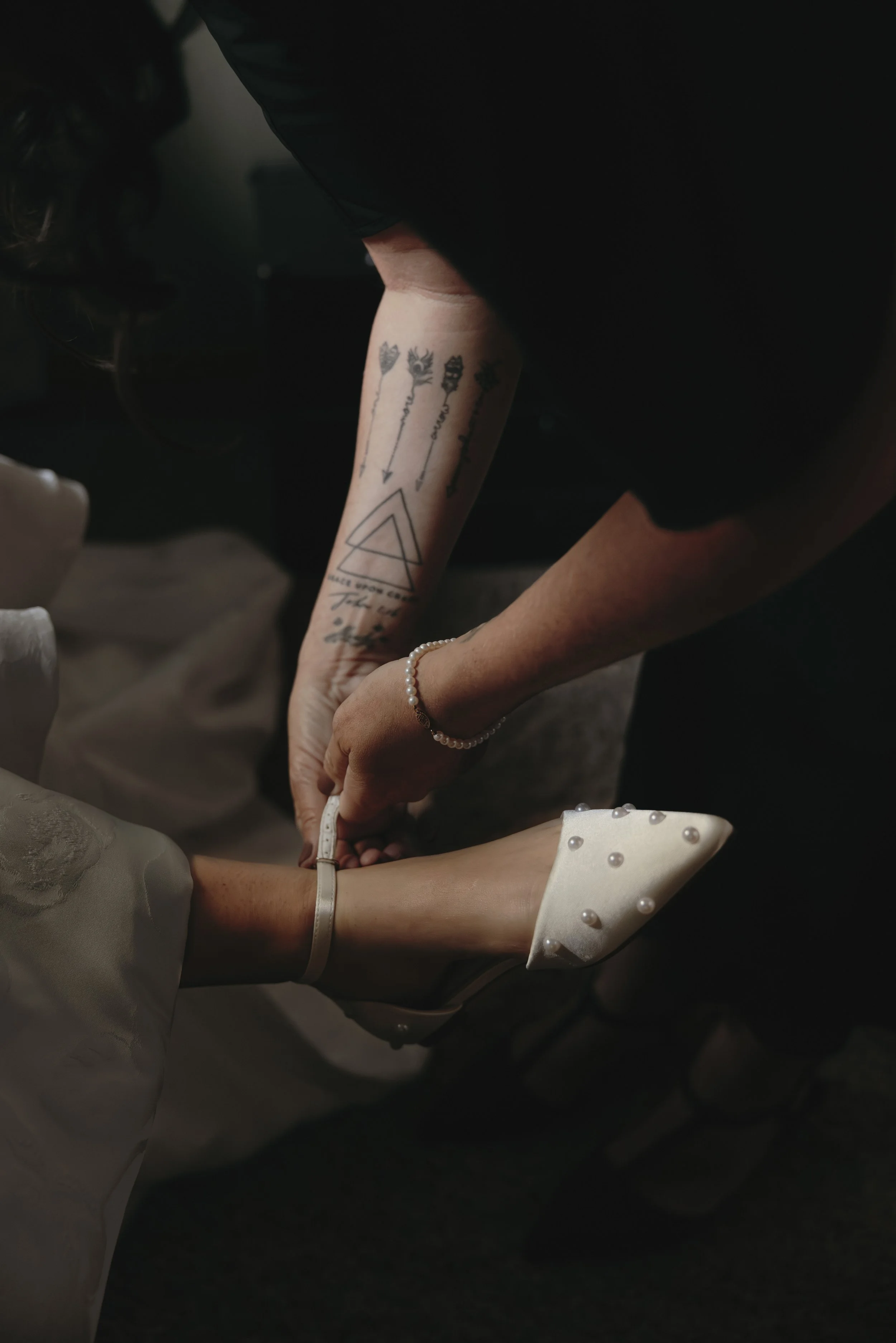 A person with tattoos on their forearm is holding the ankle of another person wearing a white dress and white high-heeled shoes with pearl embellishments.