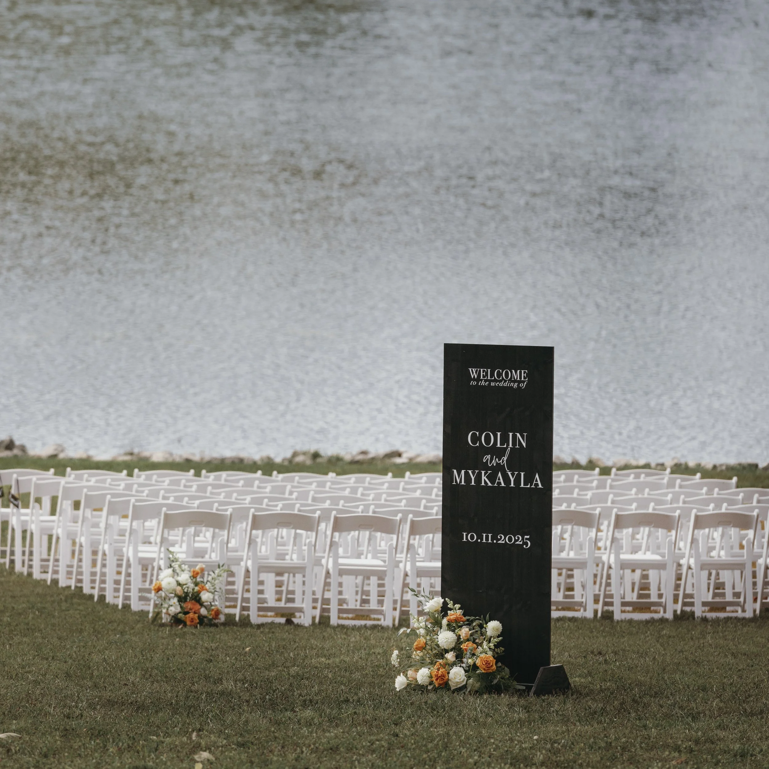 Wedding seating area by a lake with rows of white chairs, a black sign with white text welcoming guests and displaying the names 'Colin and Mykayla' and the date '10.11.2025,' decorated with flower arrangements of white, orange, and peach blooms.