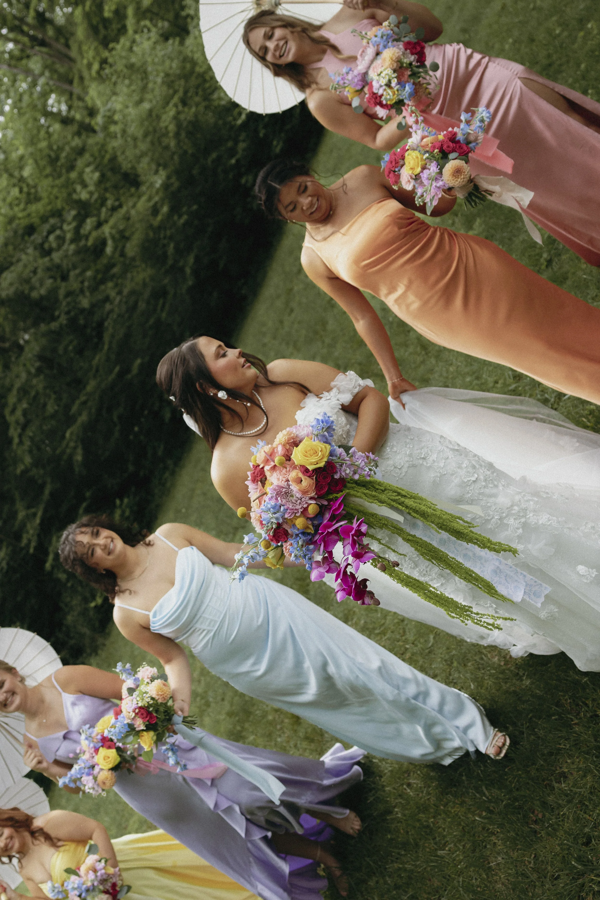 A group of women, one of whom is dressed in a white wedding gown, pose outdoors on a grassy area with green trees in the background. They are holding colorful bouquets of flowers, and some are holding white parasols.