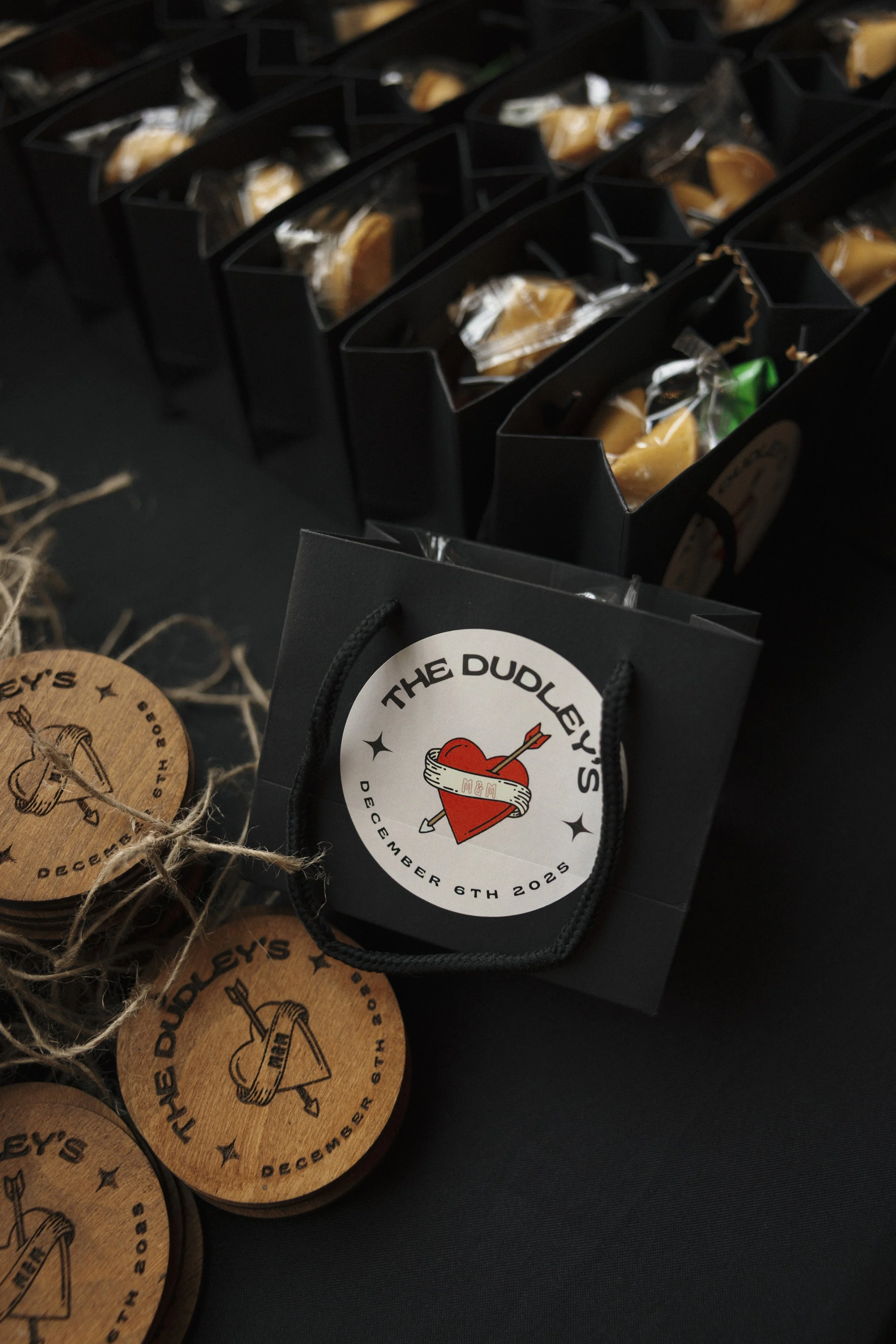 Black gift bag with a round sticker reading 'The Dudley's' and a date of December 6th, 2025, with a heart and arrow logo. Surrounding the bag are wooden coasters with the same logo and date, tied with string, and chocolate or caramel candies in black