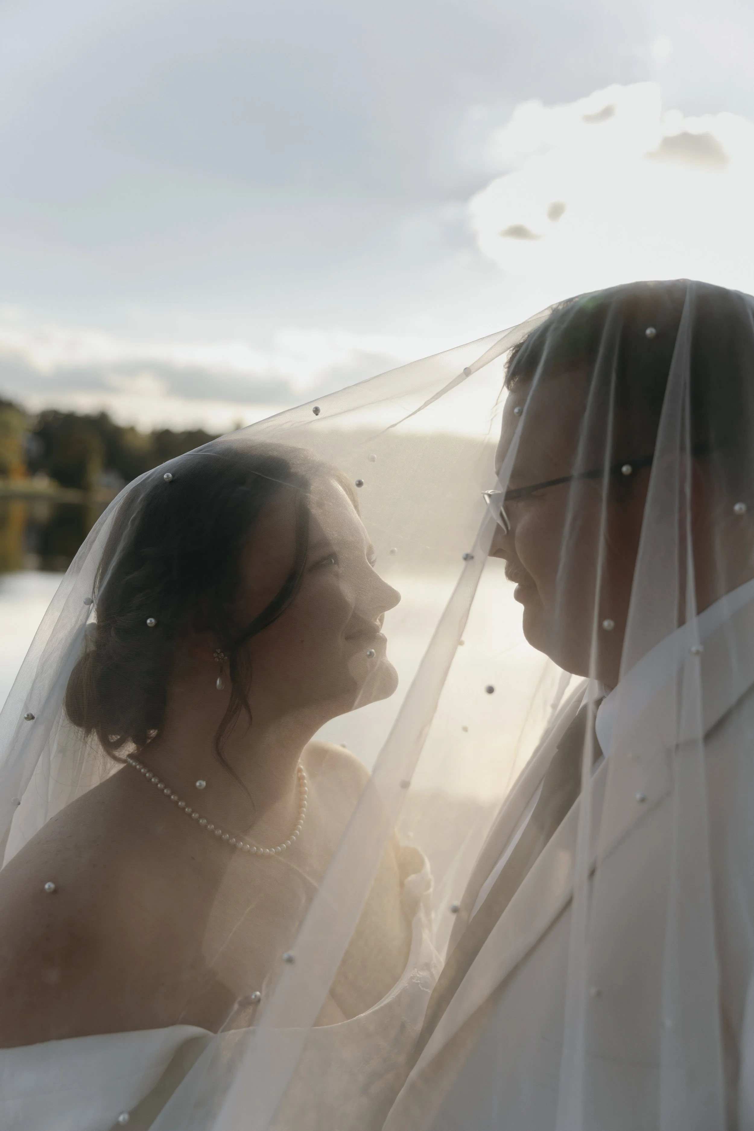 A bride and groom under a wedding veil beside a lake, facing each other at sunset.