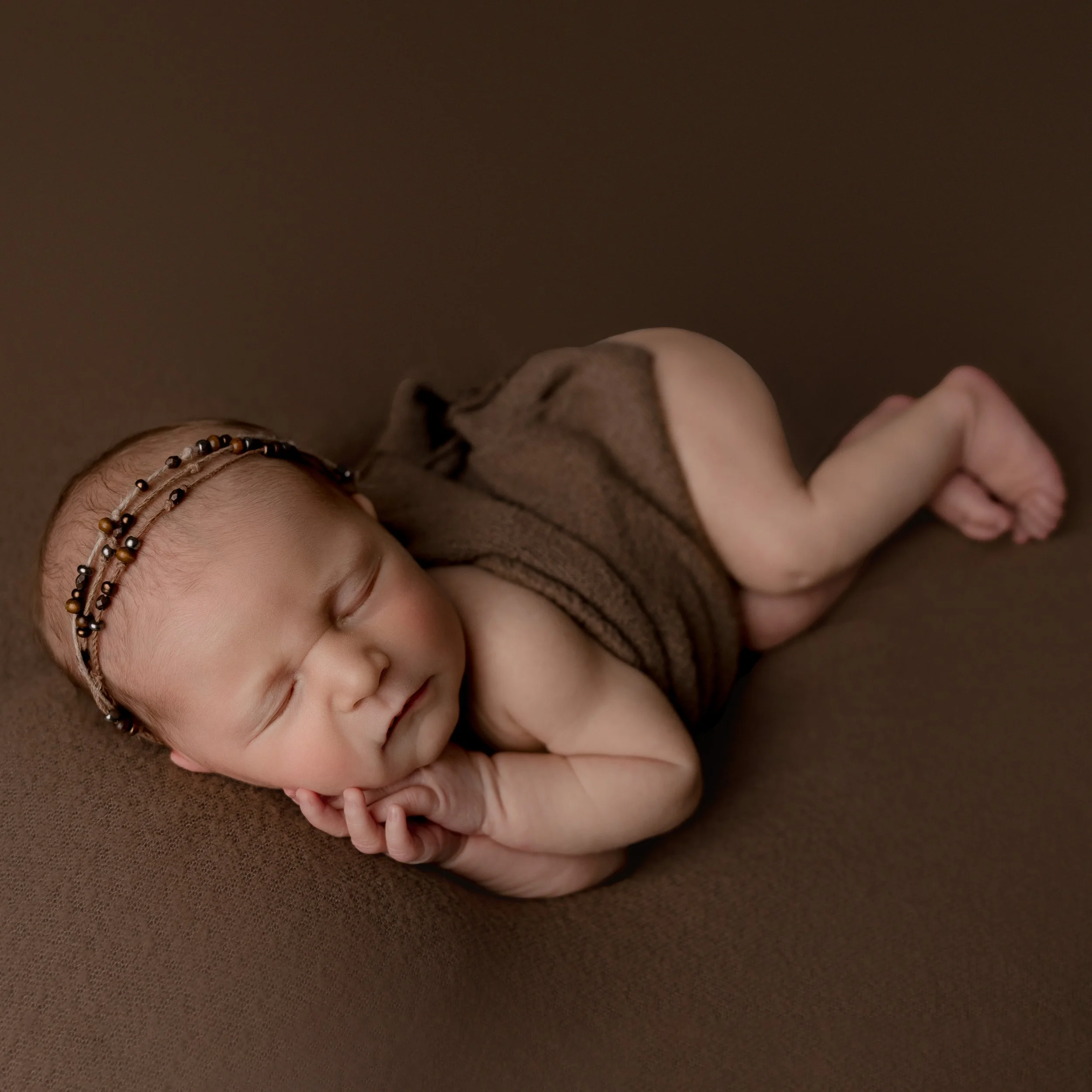 A sleeping newborn baby lying on a brown surface, wearing a brown blanket and a headband with beads.