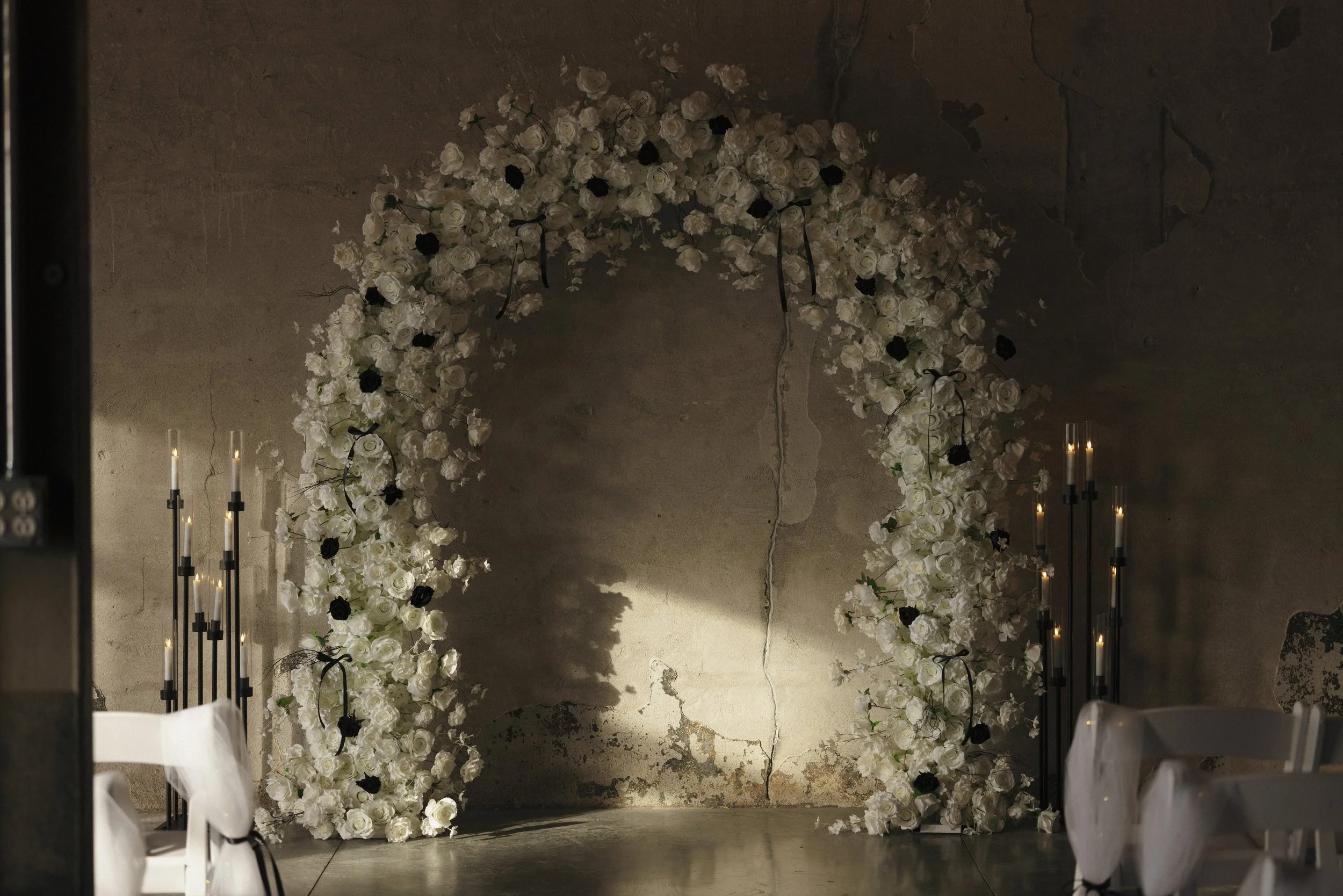 A wedding or event arch made of white and black roses, set against a rustic, cracked wall with soft lighting and candles on both sides.