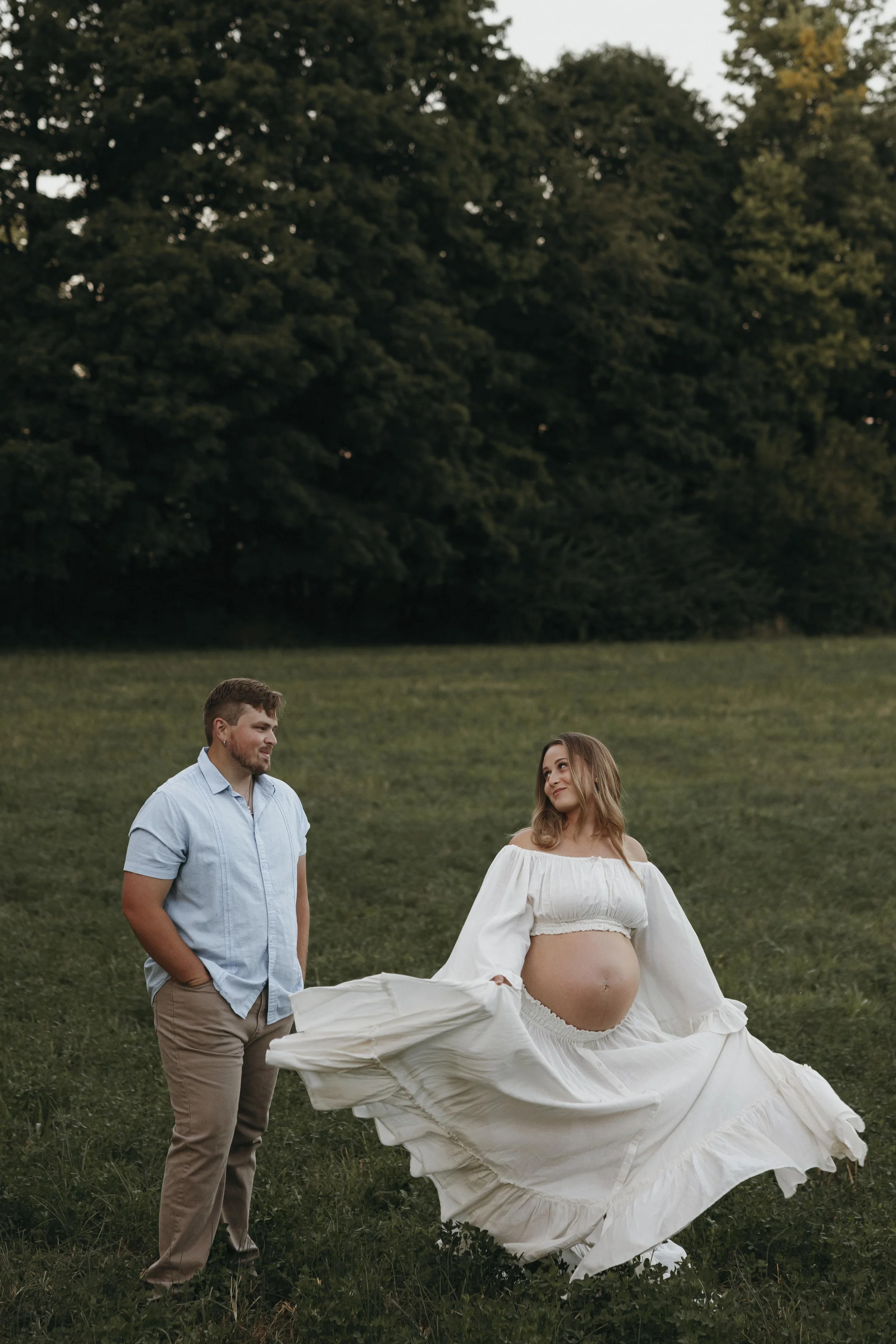 A pregnant woman in a flowing white dress stands outdoors in a grassy field holding her dress with both hands, looking at a man who is standing nearby with his hands in his pockets, both smiling softly with trees in the background.