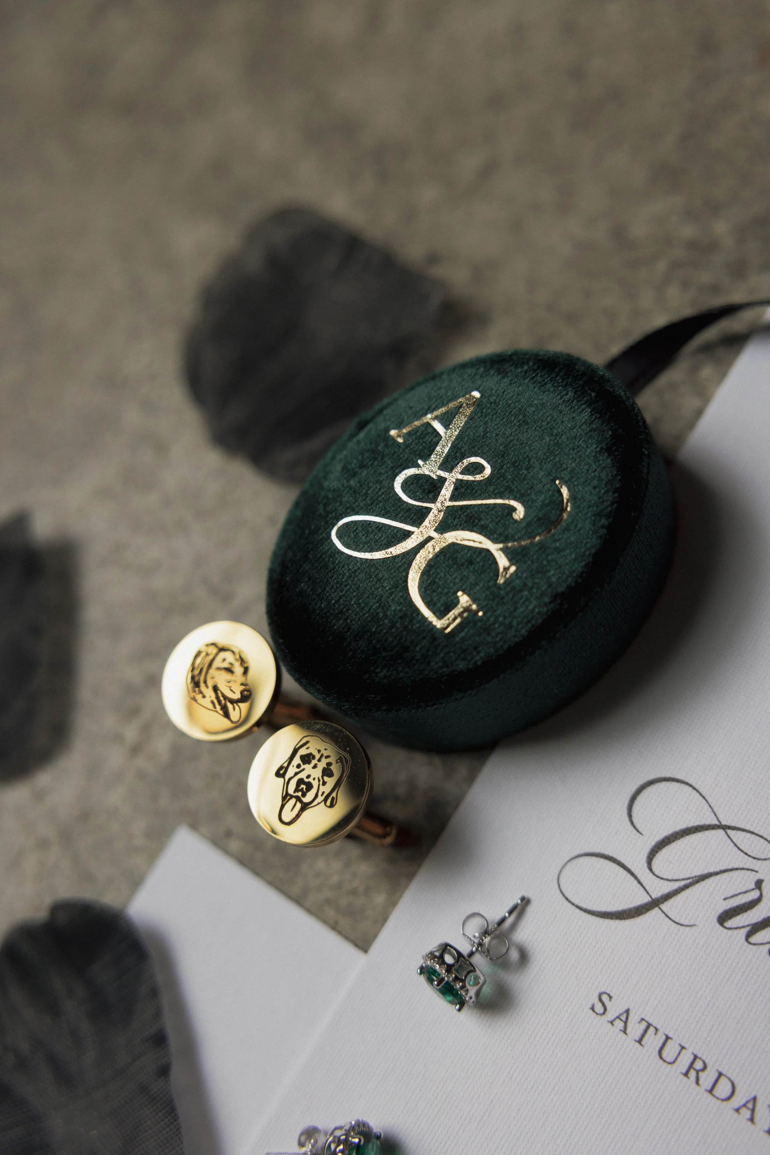Close-up of a green circular box with gold logo initials 'A & G'. Two pairs of earrings depict dog and woman caricatures, laid on a table with some paper and rocks in the background.