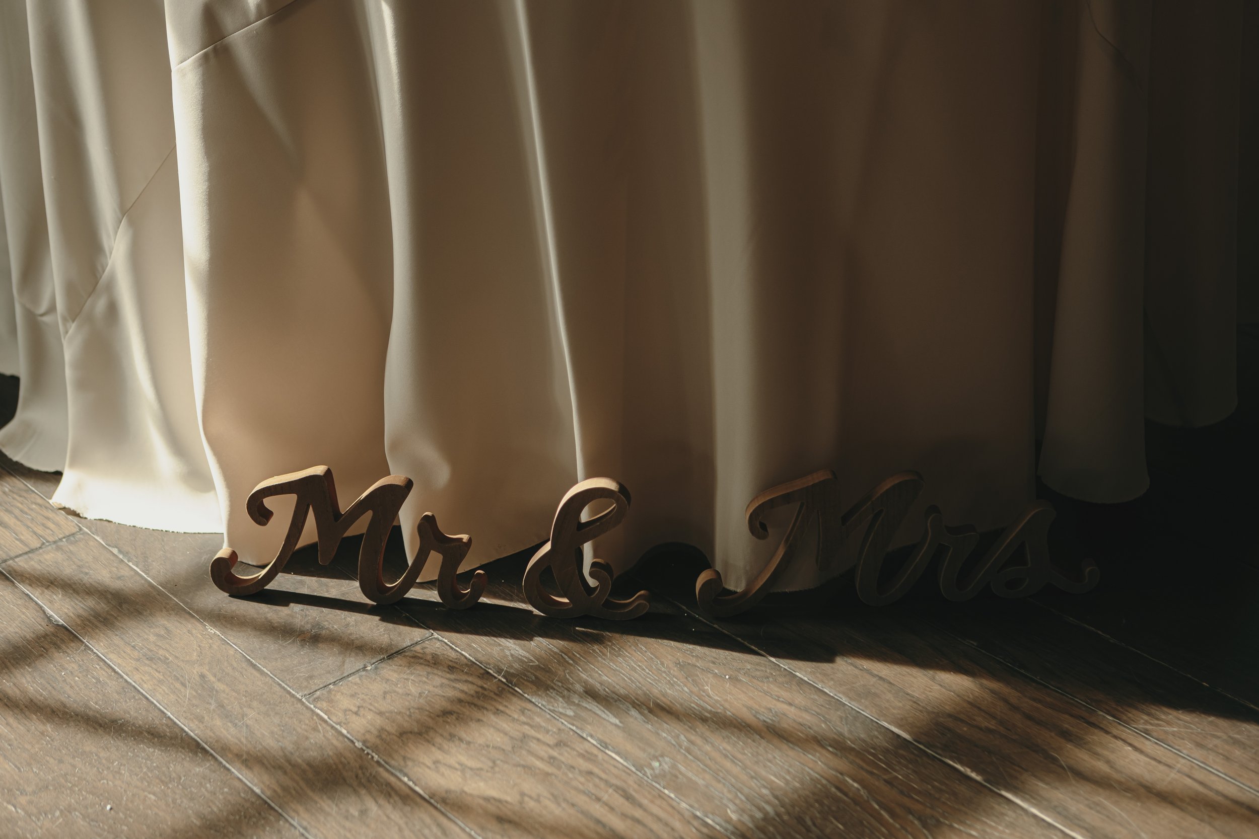 Wooden 'Mr & Mrs' sign on a wooden floor next to a cream-colored curtain.