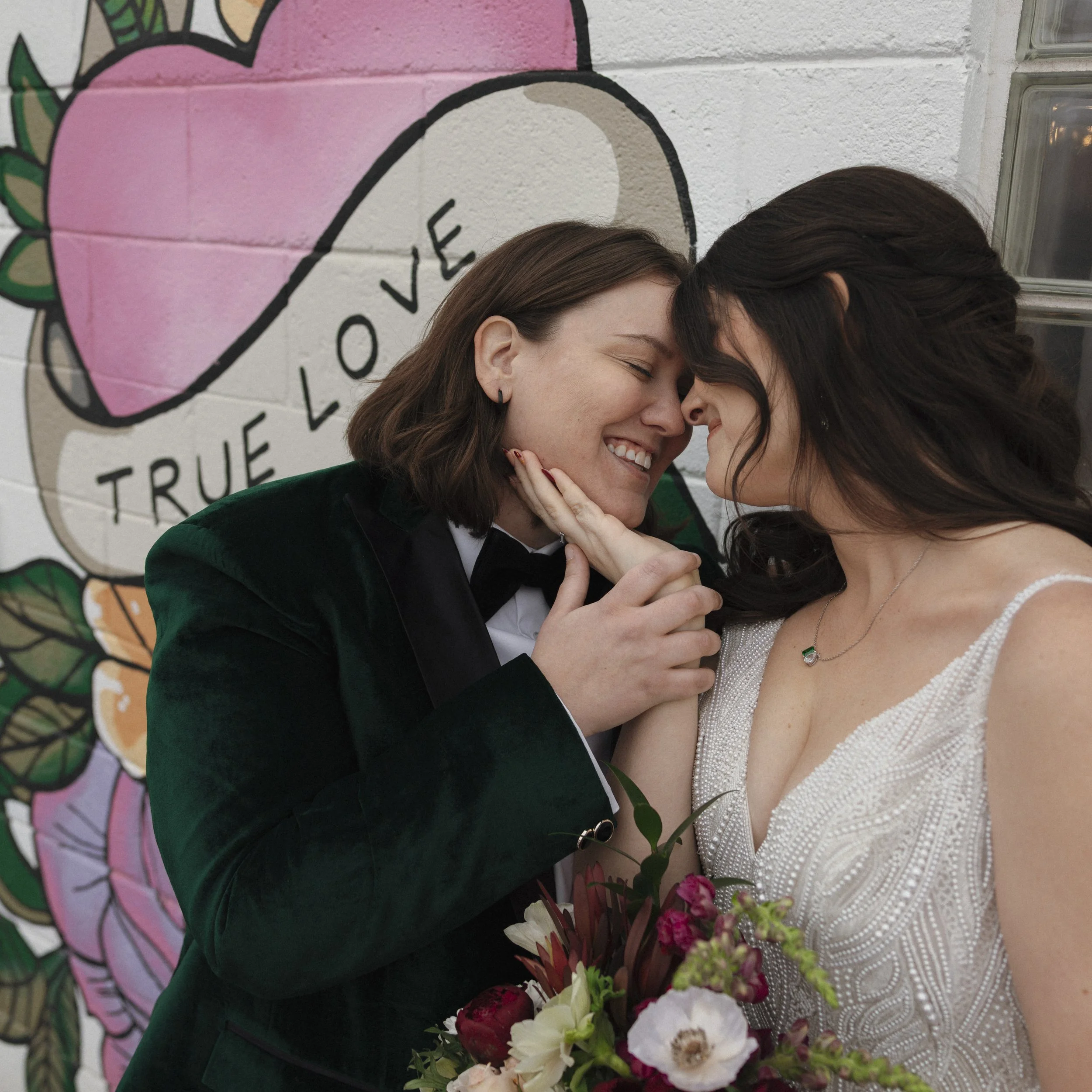 A couple sharing an intimate moment at their wedding, with their foreheads touching and eyes closed, holding a bouquet of flowers. They are standing in front of a colorful mural with the words 'True Love' written on it.