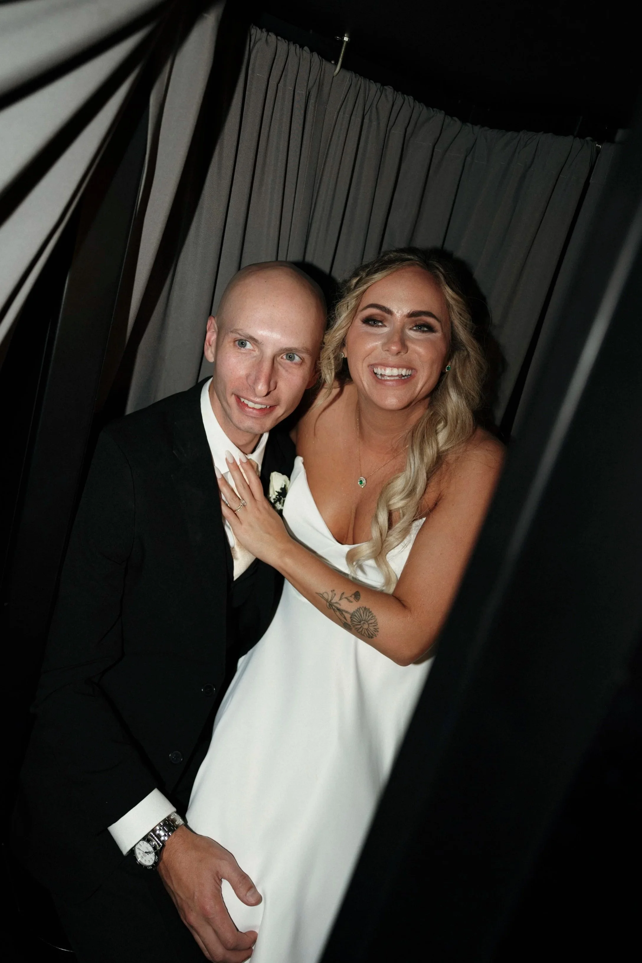 A bride and groom smiling inside a photo booth at their wedding, with dark curtains in the background.