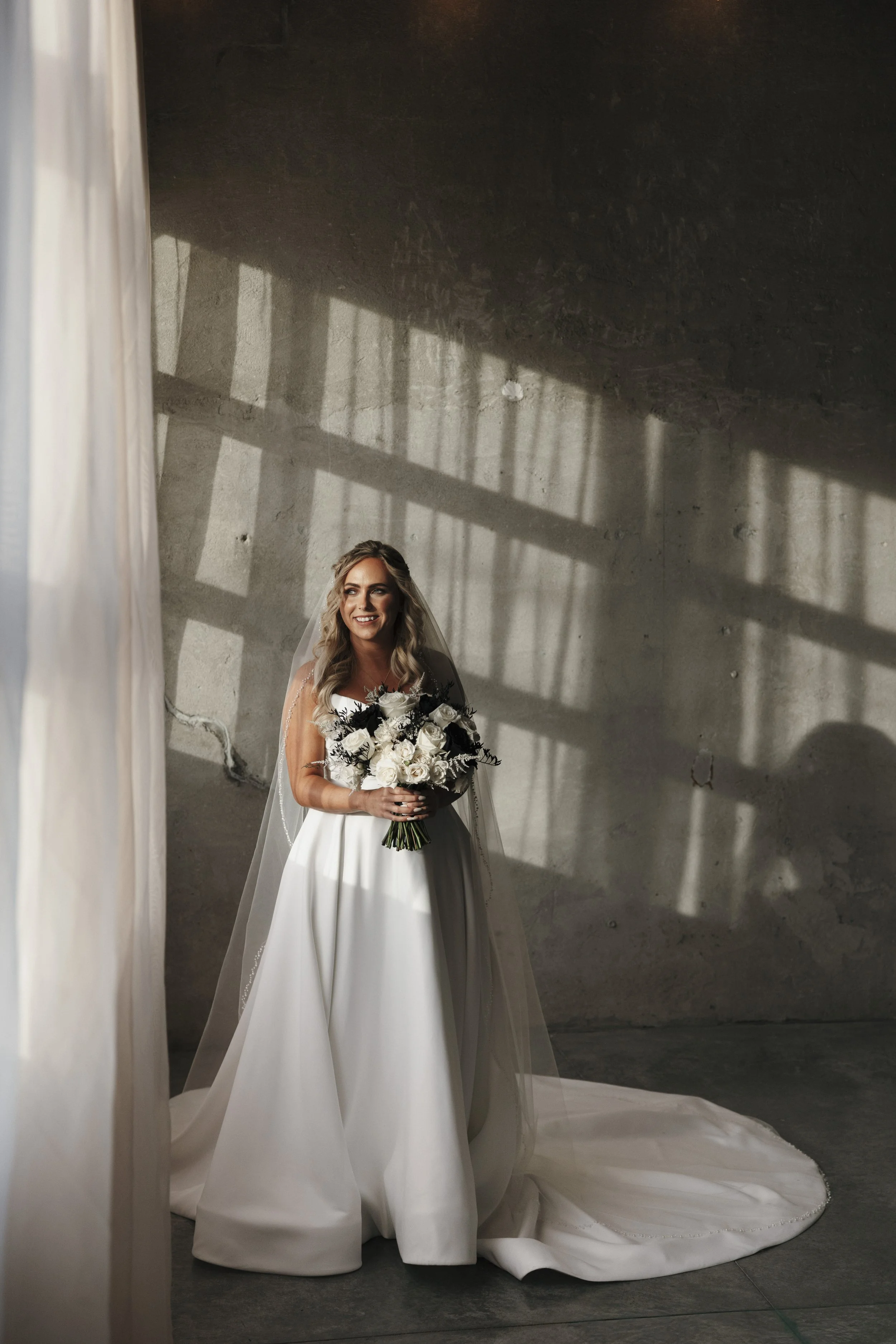 A bride stands in a minimalist industrial space, smiling and holding a bouquet of white and dark flowers, with sunlight casting shadows on the concrete wall.