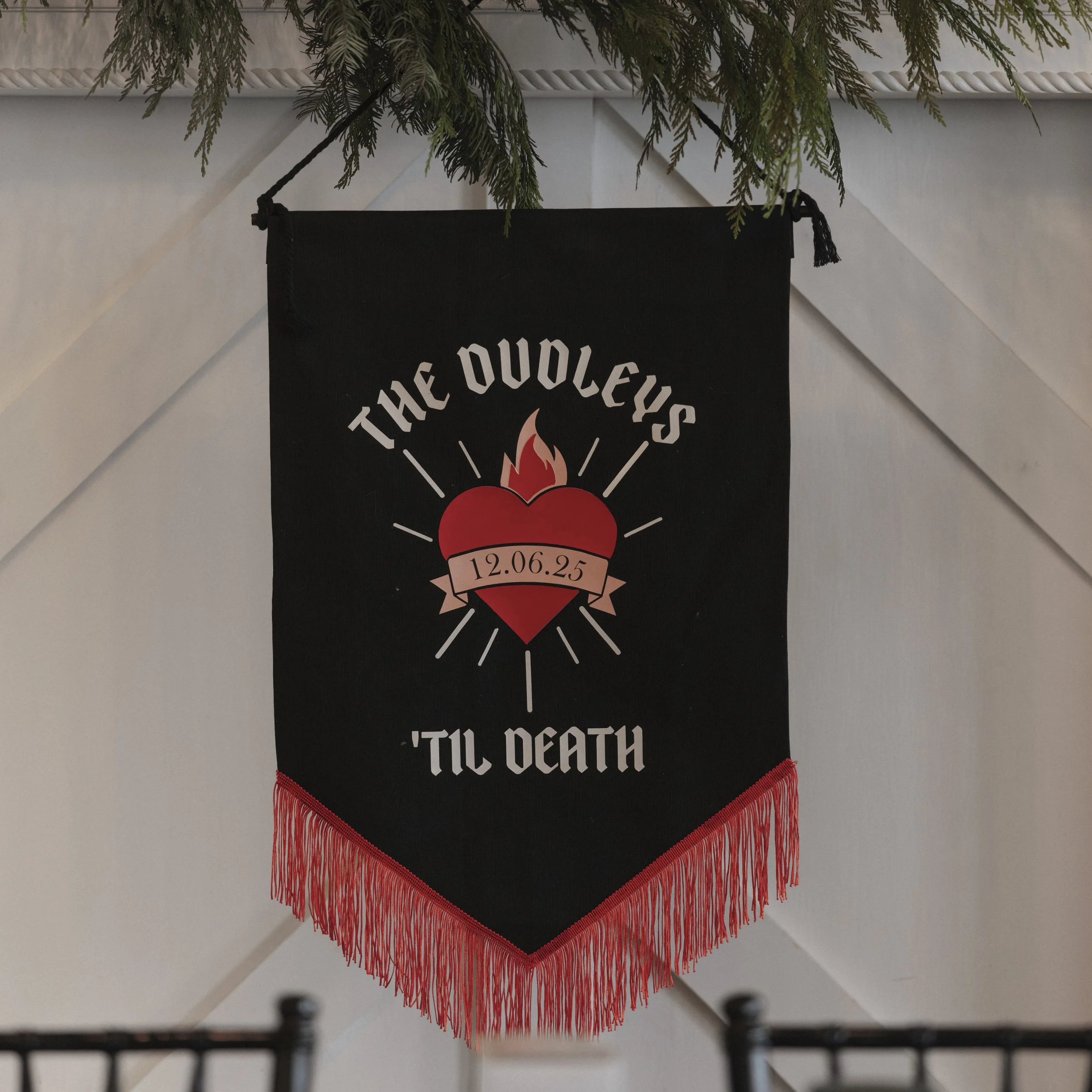 Black banner with red heart and flames, white text reads 'The Doodleys' and 'Til Death,' with a pink banner displaying the date '12.06.25,' and red fringe at the bottom.