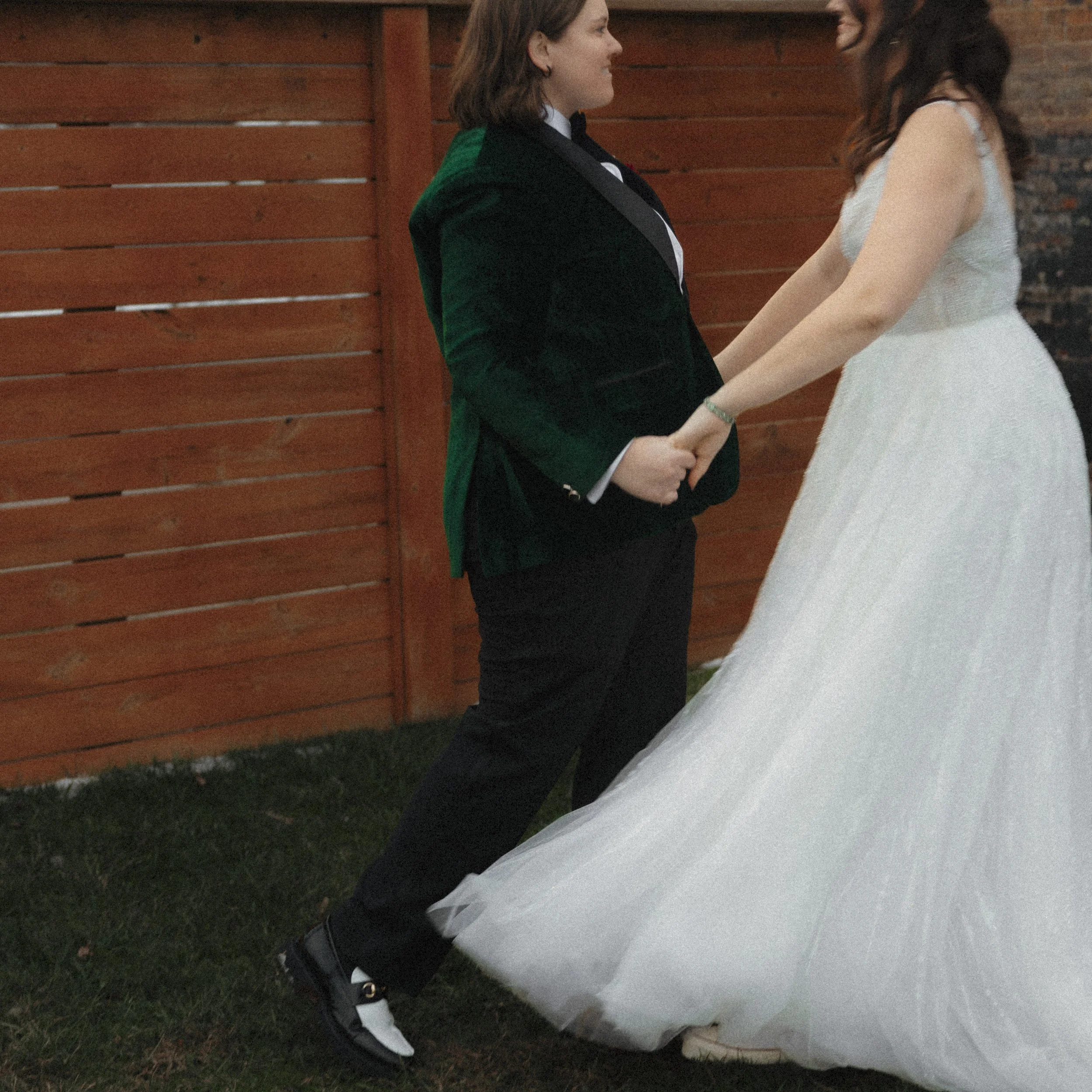 A couple holding hands and facing each other in front of a wooden fence, with the woman in a wedding dress and the man in a tuxedo.