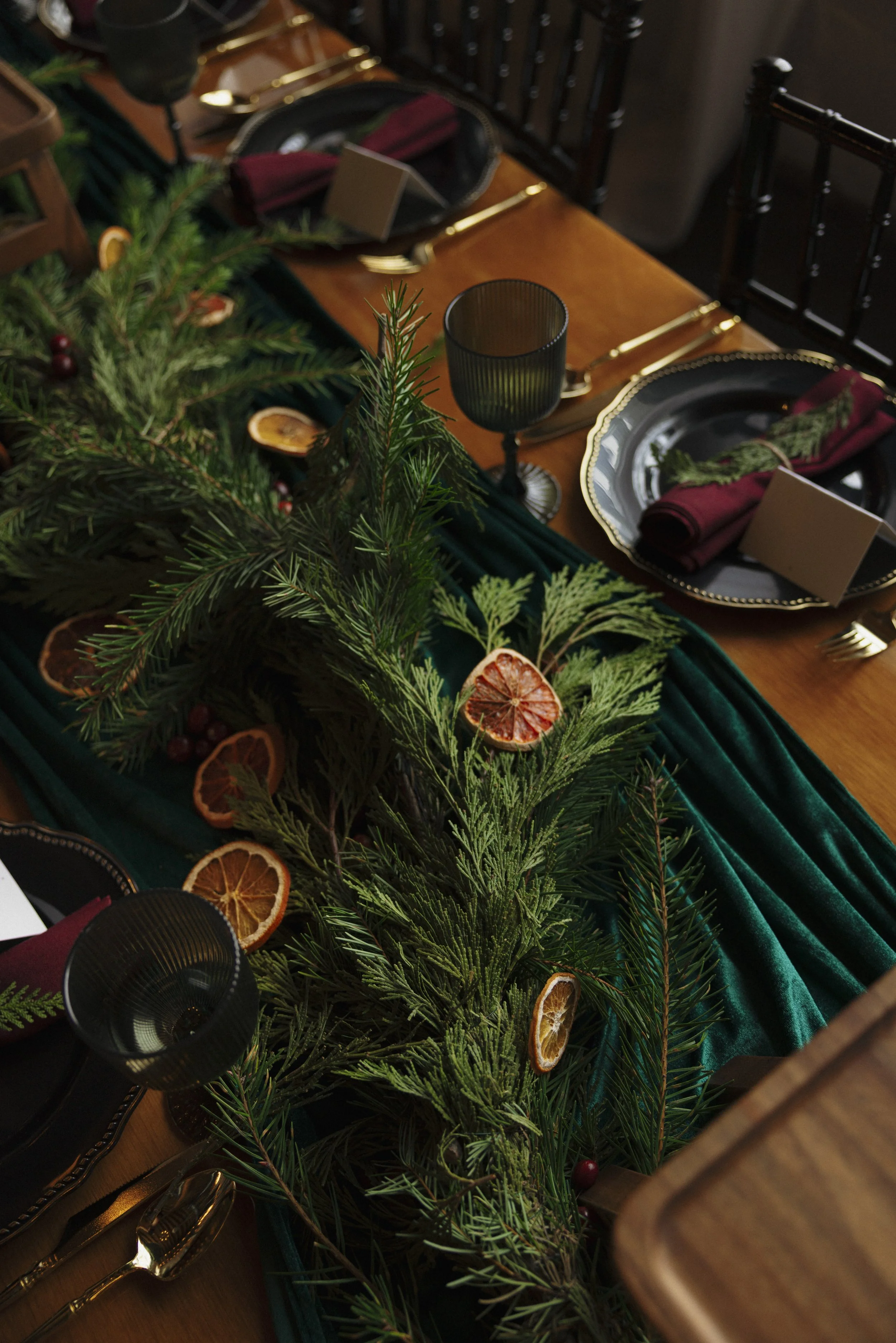 A festive holiday dining table decorated with evergreen branches and dried orange slices as a centerpiece, with black and gold plates, burgundy napkins, gold flatware, black glasses, and place cards.